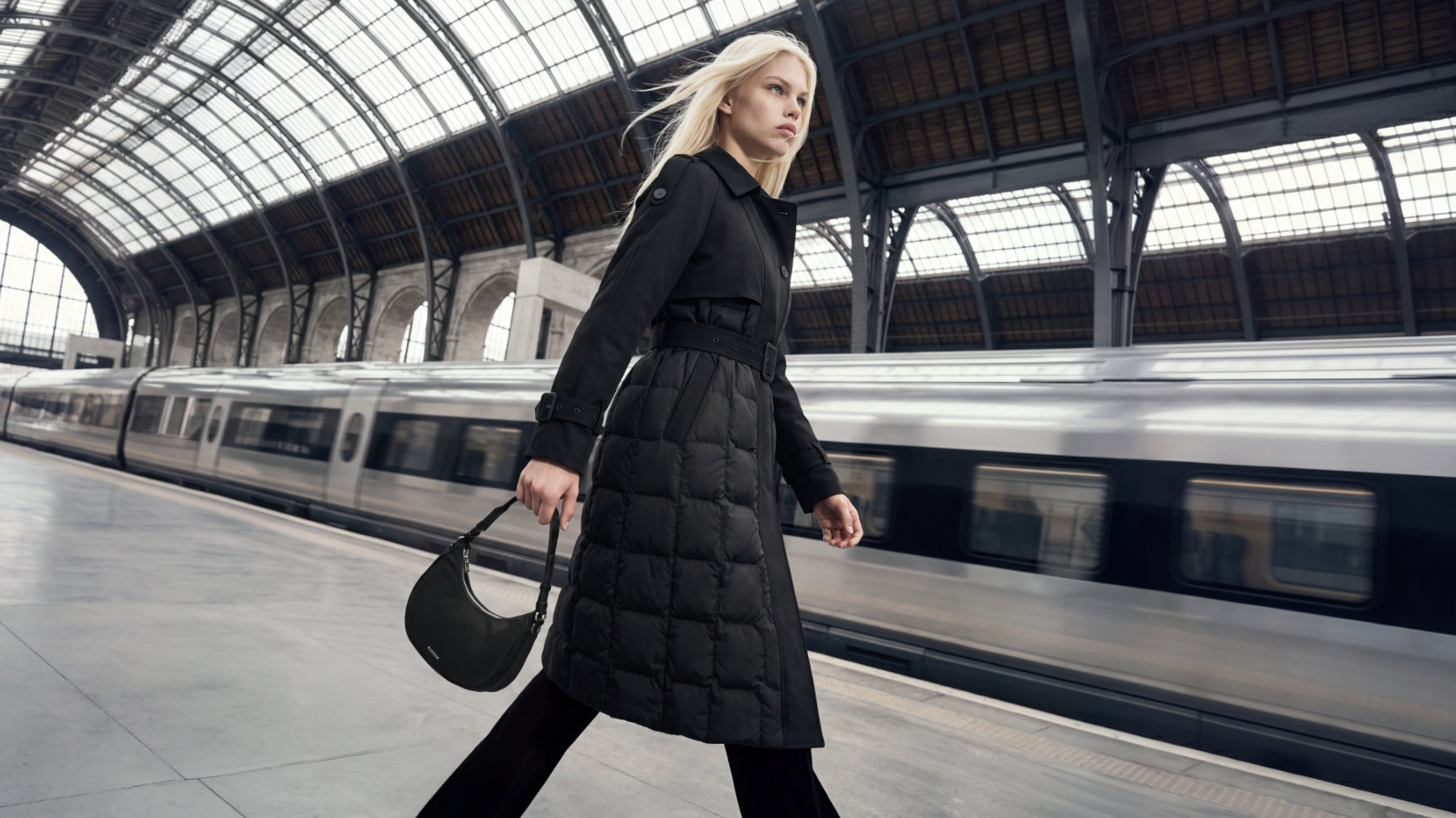 A woman walking on a train station platform carrying a black shoulder bag, wearing a long black quilted puffer coat and black pants, with a silver train and a large arched glass station hall in the background.