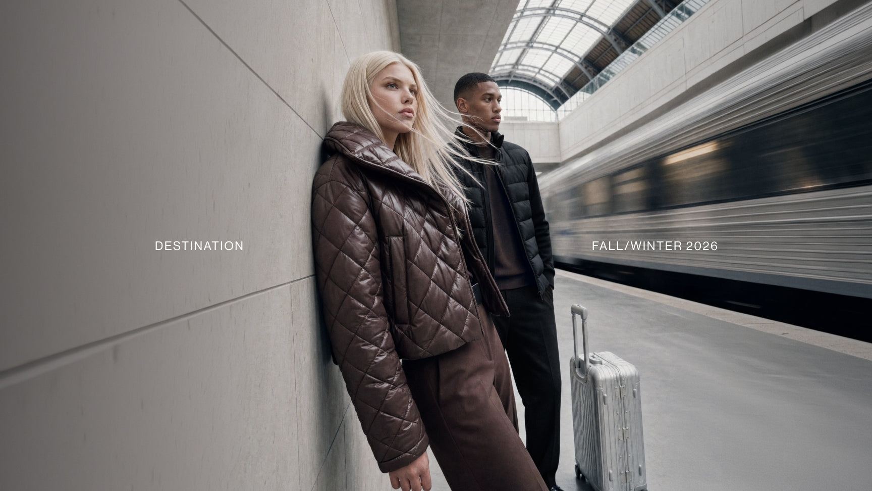 A blonde woman in a brown quilted puffer jacket leans against a concrete wall at a train station platform, with a man in a black puffer jacket standing behind her. A silver suitcase sits on the platform, and a train passes in the blurred background.