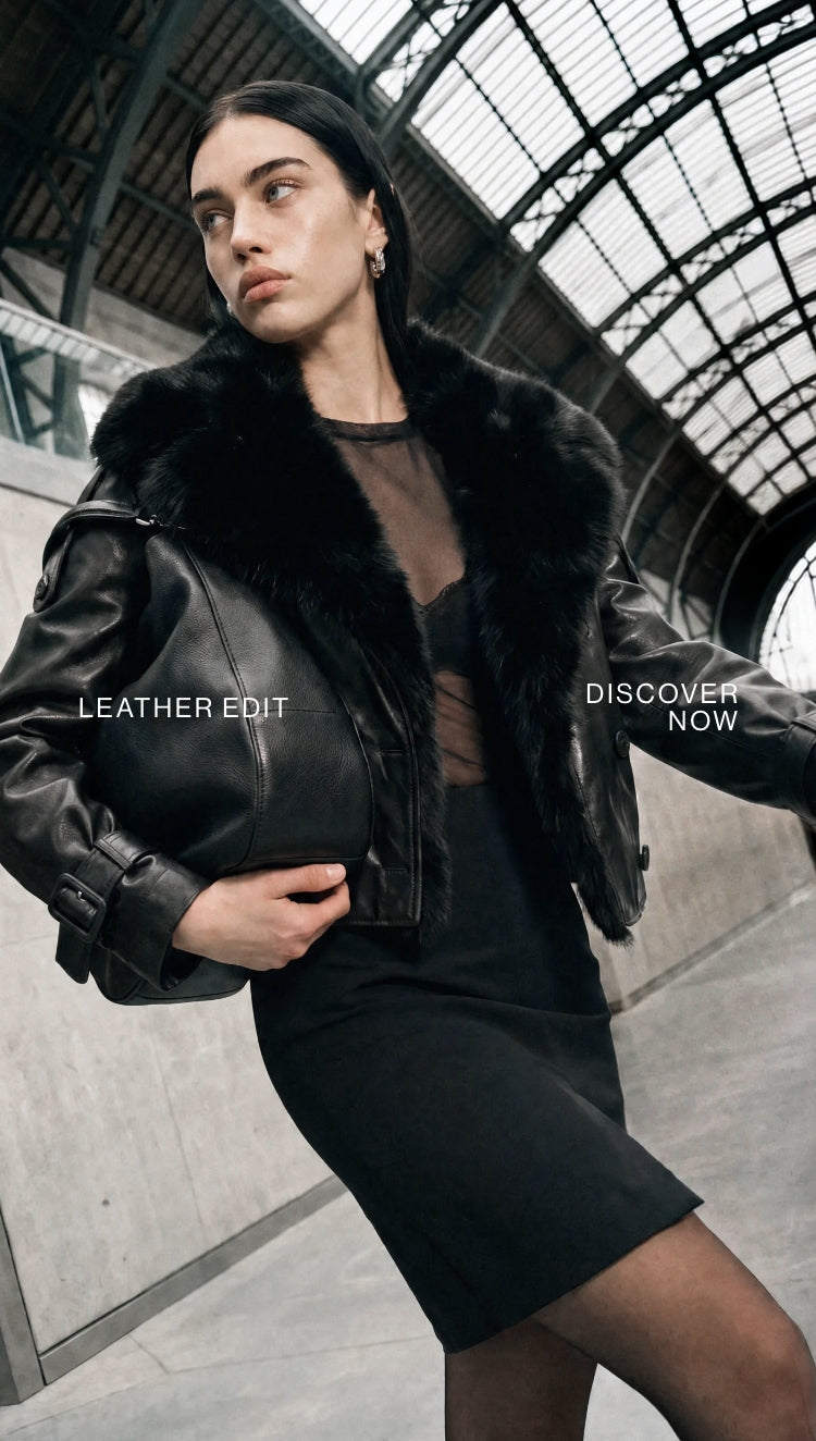 A woman in a black leather jacket with a black fur collar stands in a large, arched train station hall, holding a black leather handbag. 