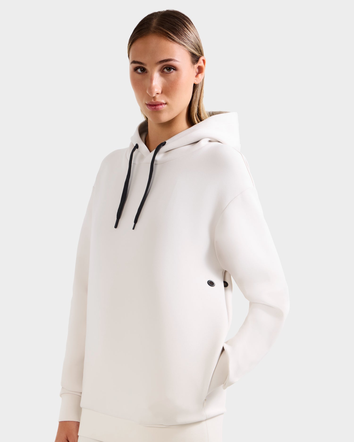 A woman with straight, light brown hair wears the Rudsak LEROY - U525509 Iceland W premium white hoodie with black drawstrings, standing against a plain light gray background.