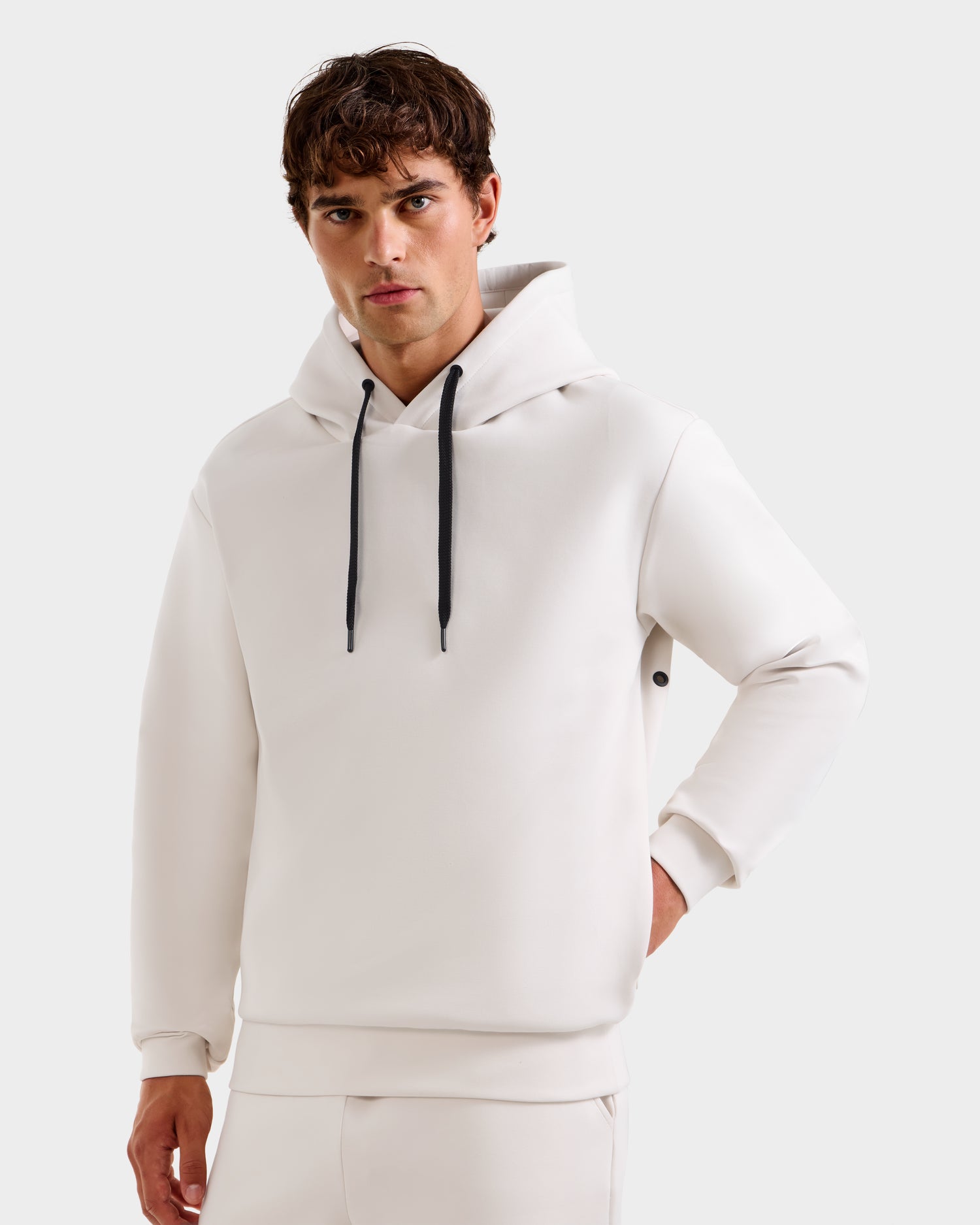 A young man with short brown hair models the Rudsak LEROY - U525509 Iceland W premium hoodie with black drawstrings and matching white pants, posing with one hand in his pocket against a plain light background.
