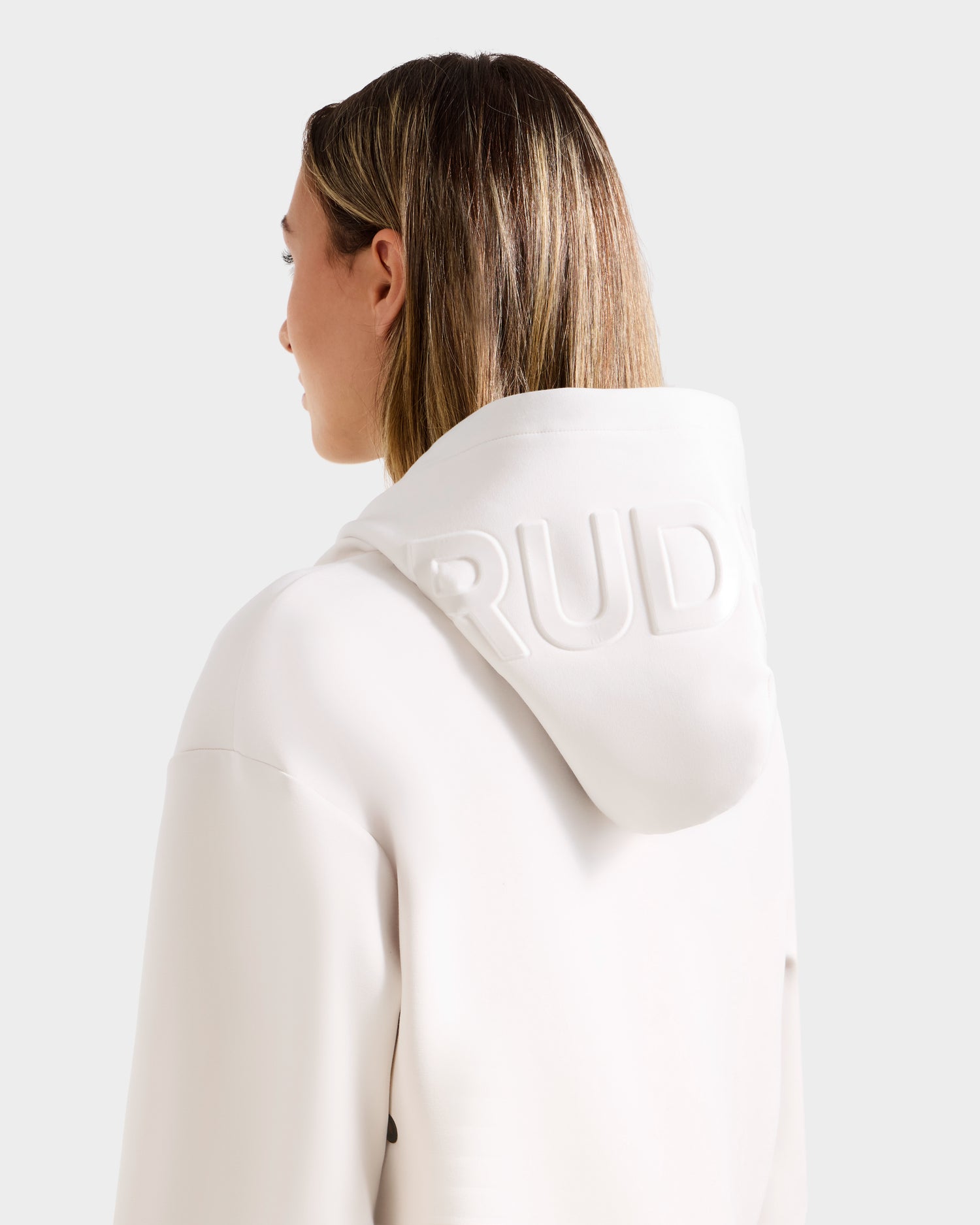 A person with straight, shoulder-length hair is seen from behind, wearing the Rudsak LEROY - U525509 Iceland W white premium unisex hoodie with "RUD" embossed on the hood against a plain light background.