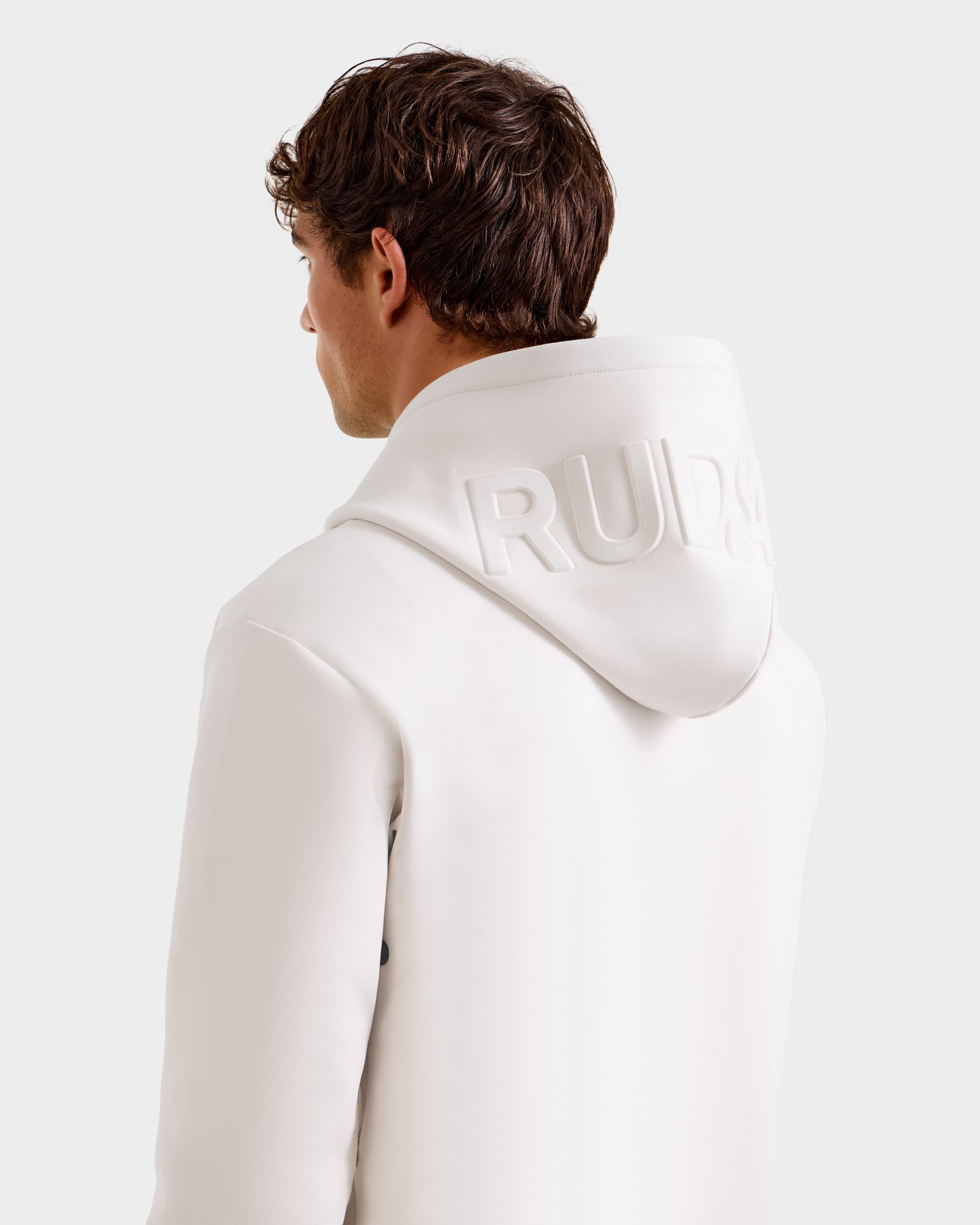 A person with short brown hair is seen from behind, wearing the Rudsak LEROY - U525509 Iceland W premium unisex hoodie. The hood features branding, and the background is plain and light-colored.