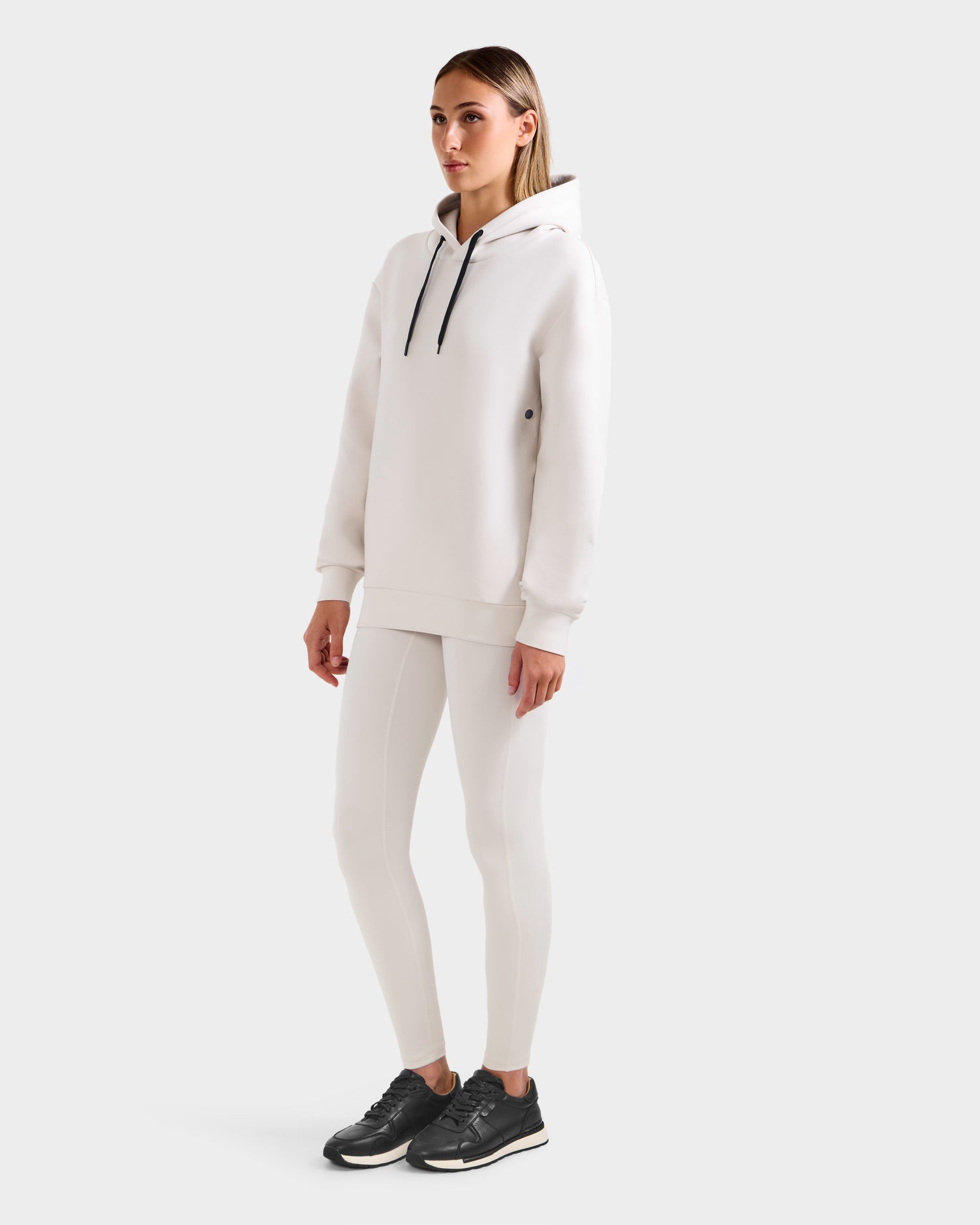 A woman stands against a plain background wearing the Rudsak LEROY - U525509 Iceland W hoodie, paired with white leggings and black athletic shoes. She gazes forward neutrally, hands relaxed at her sides.