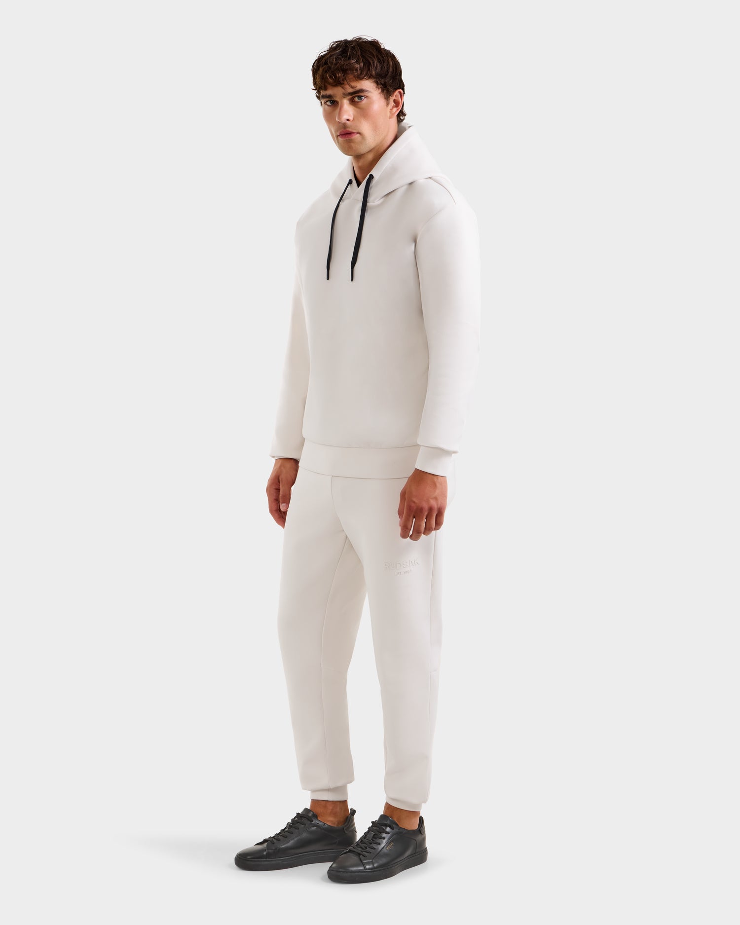 A man models the Rudsak LEROY - U525509 Iceland M premium, gender-inclusive hoodie with black drawstrings and matching white jogger pants, paired with black sneakers, facing slightly to the side against a plain white background.