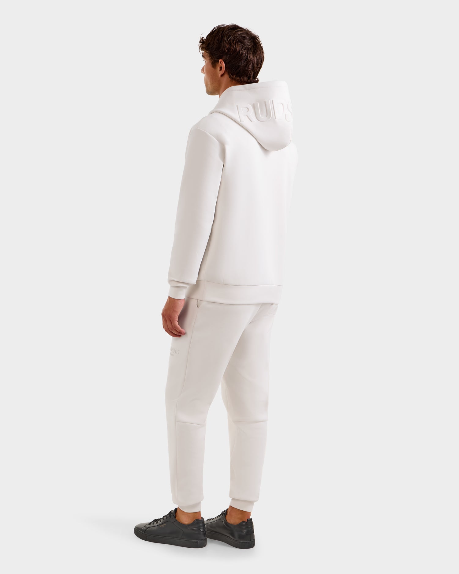 A person faces away, wearing the LEROY - U525509 Iceland M by Rudsak: a premium, all-white gender-inclusive hoodie and sweatpants set with embossed hood text and black sneakers, against a plain white background.