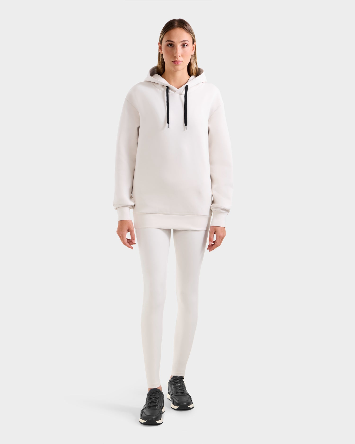 A person stands facing forward, wearing the Rudsak LEROY - U525509 Iceland W premium unisex hoodie with black drawstrings, white leggings, and black athletic shoes against a plain light background.