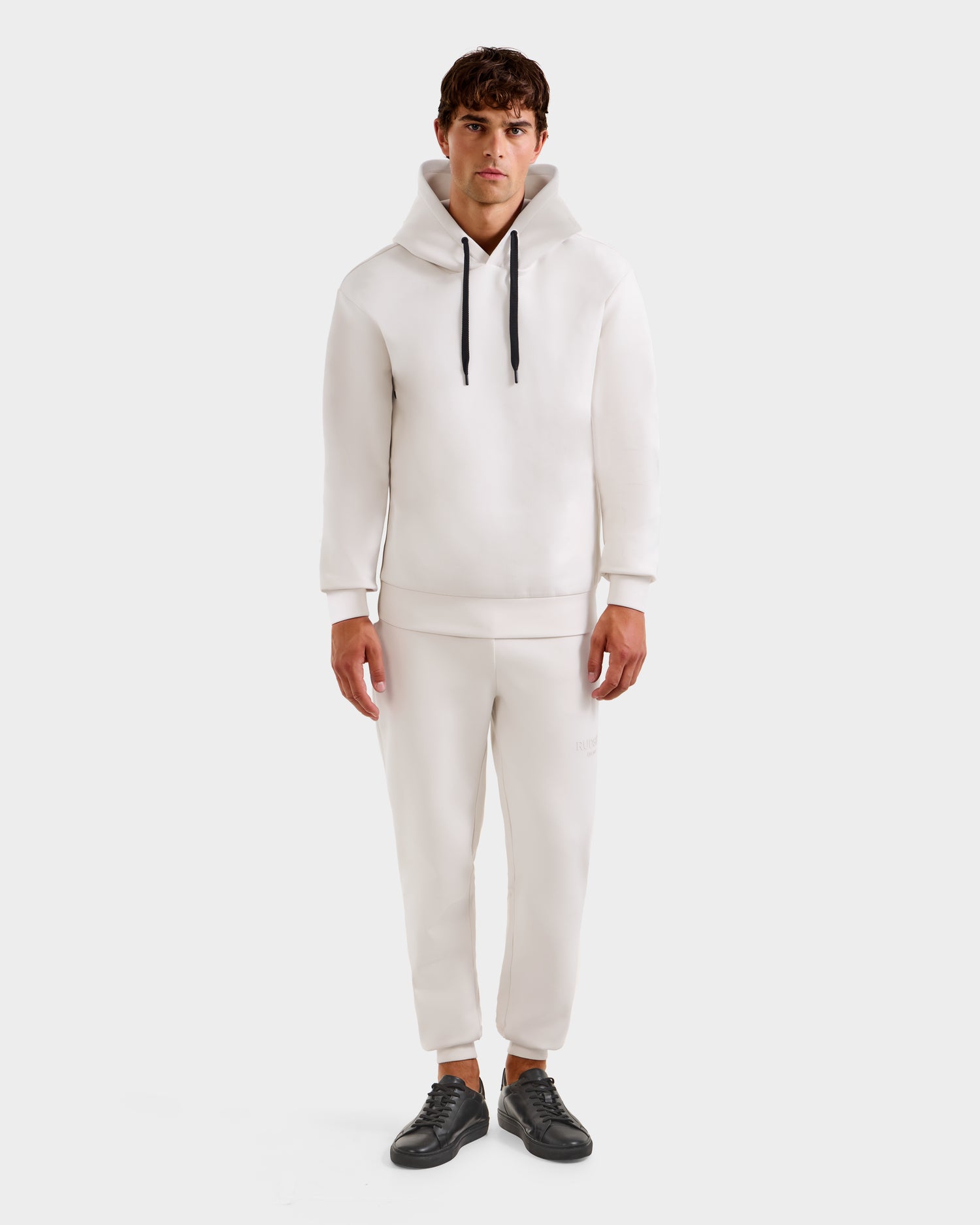 A man faces forward, dressed in the LEROY - U525509 Iceland M hoodie by Rudsak with black drawstrings, paired with white jogger pants and black sneakers. The plain white background highlights the outfit.