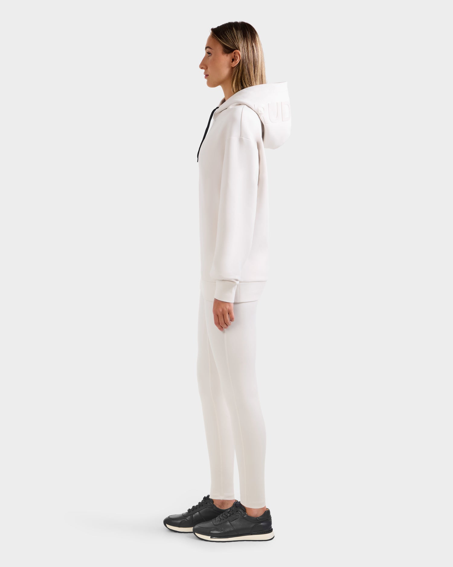 A person stands in profile wearing the Rudsak LEROY - U525509 Iceland W hoodie and matching leggings with black sneakers, set against a plain white background.