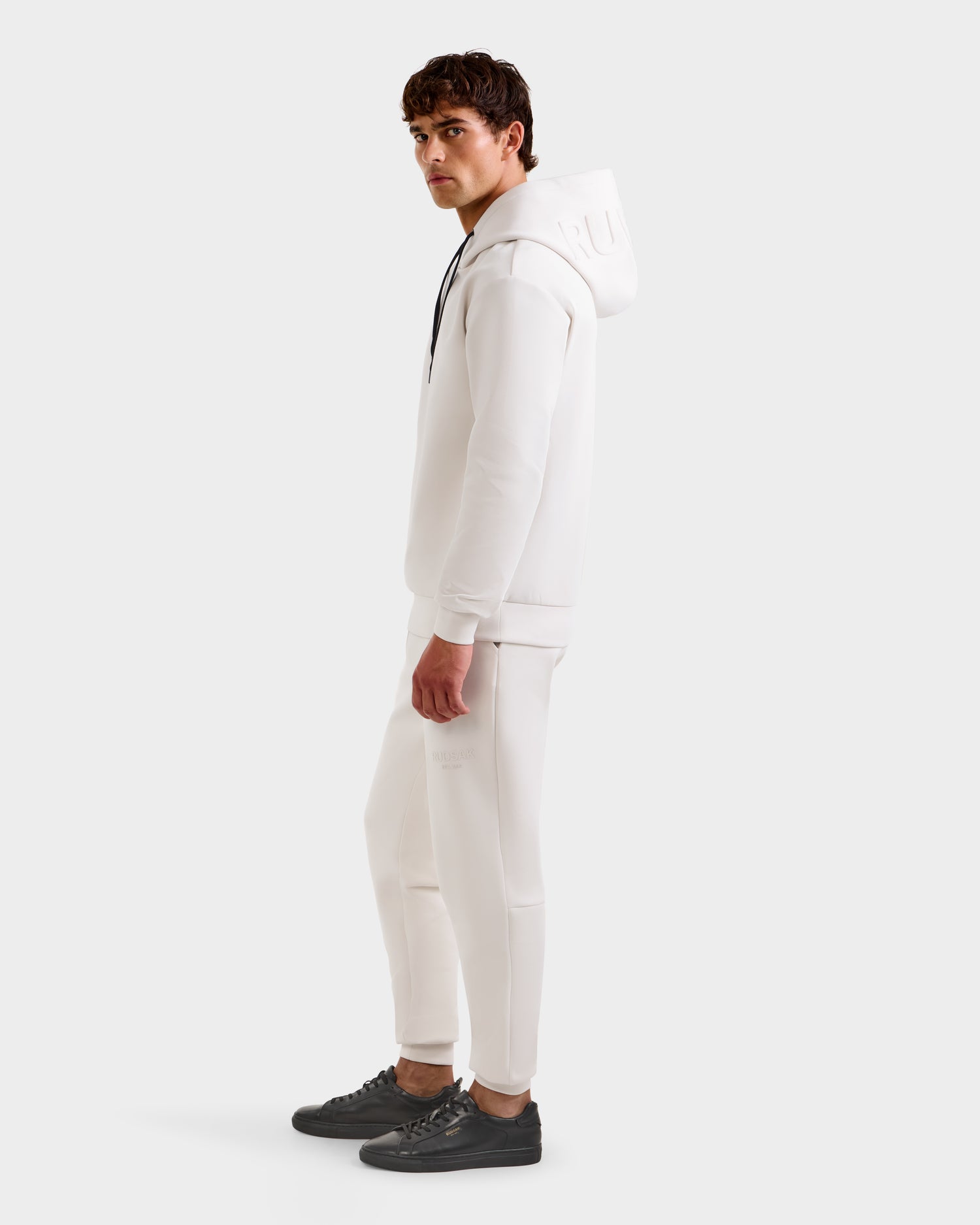 A man stands sideways wearing the Rudsak LEROY - U525509 Iceland M hoodie with matching white joggers and black sneakers, set against a plain white background.