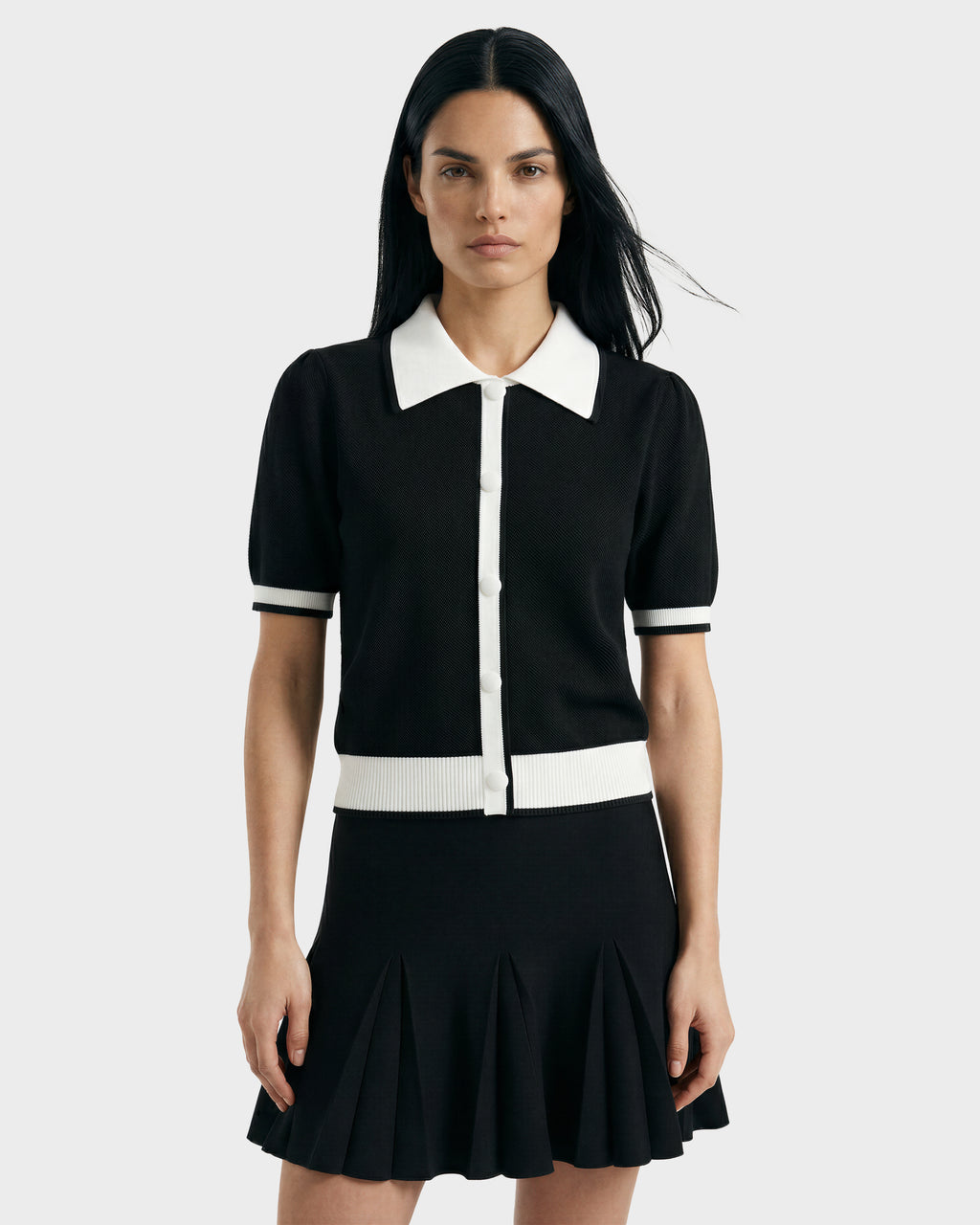 A woman with long dark hair models the IRIS - 8526012 Black short-sleeve sweater with white trim and buttons by Rudsak, paired with a black pleated skirt against a plain light gray background.
