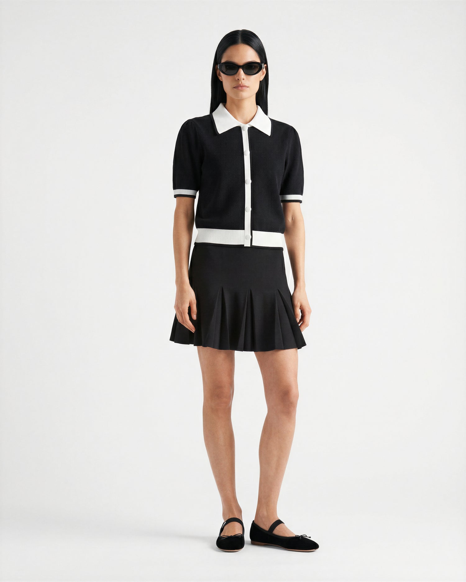 A person stands against a white background wearing Rudsak's IRIS - 8526012 Black sunglasses, a knit short-sleeve sweater with contrast collar and trim, a black pleated mini skirt, and black slip-on shoes.