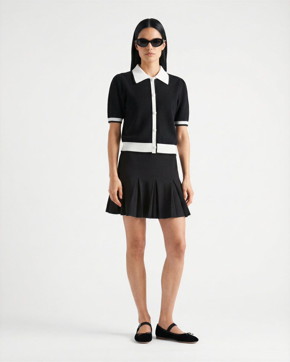 A person stands against a white background wearing Rudsak's IRIS - 8526012 Black sunglasses, a knit short-sleeve sweater with contrast collar and trim, a black pleated mini skirt, and black slip-on shoes.