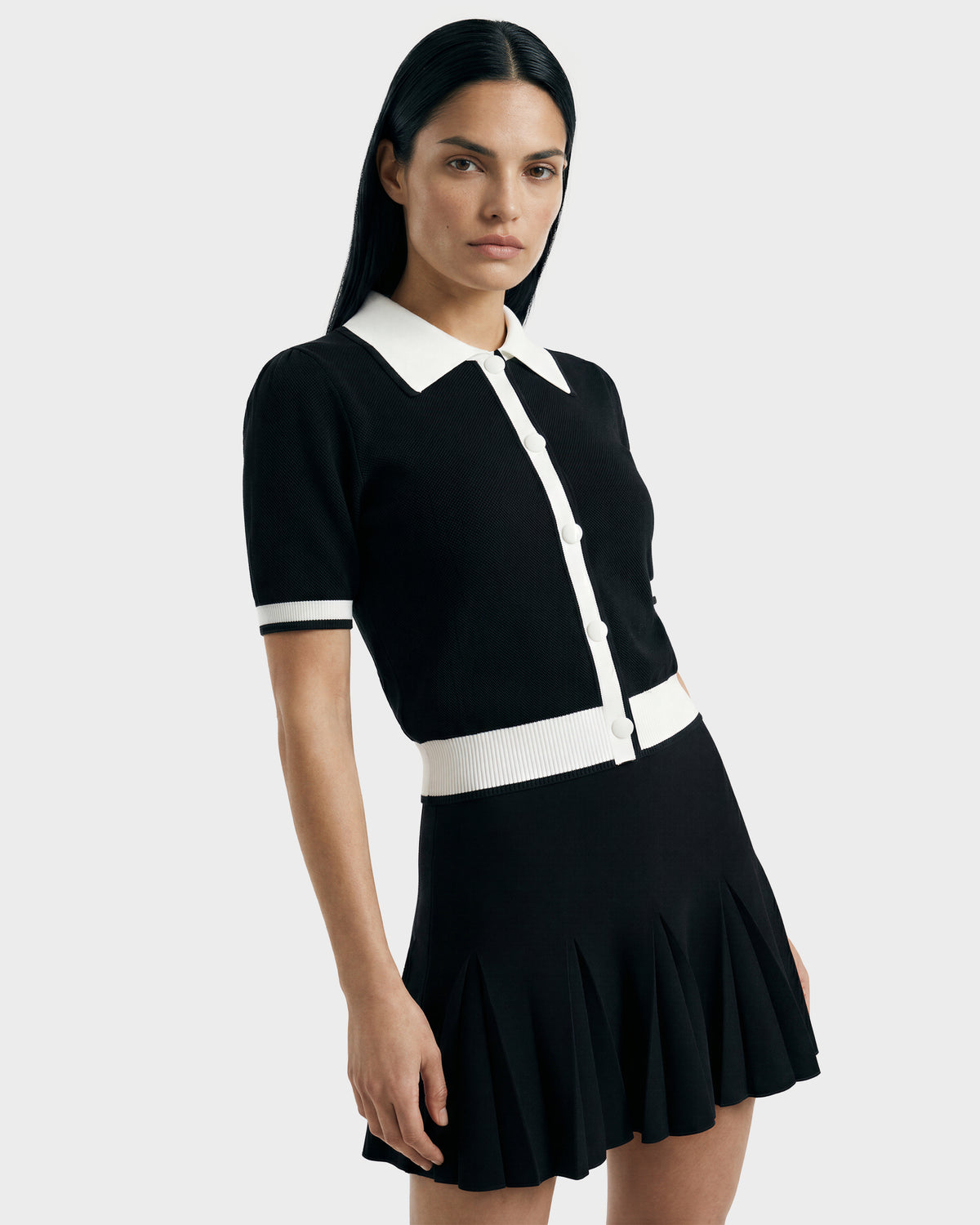 A woman with straight dark hair wears the Rudsak IRIS - 8526012 Black knit short-sleeve sweater with a white contrast collar, buttons, and trim, paired with a black pleated skirt against a plain light background.