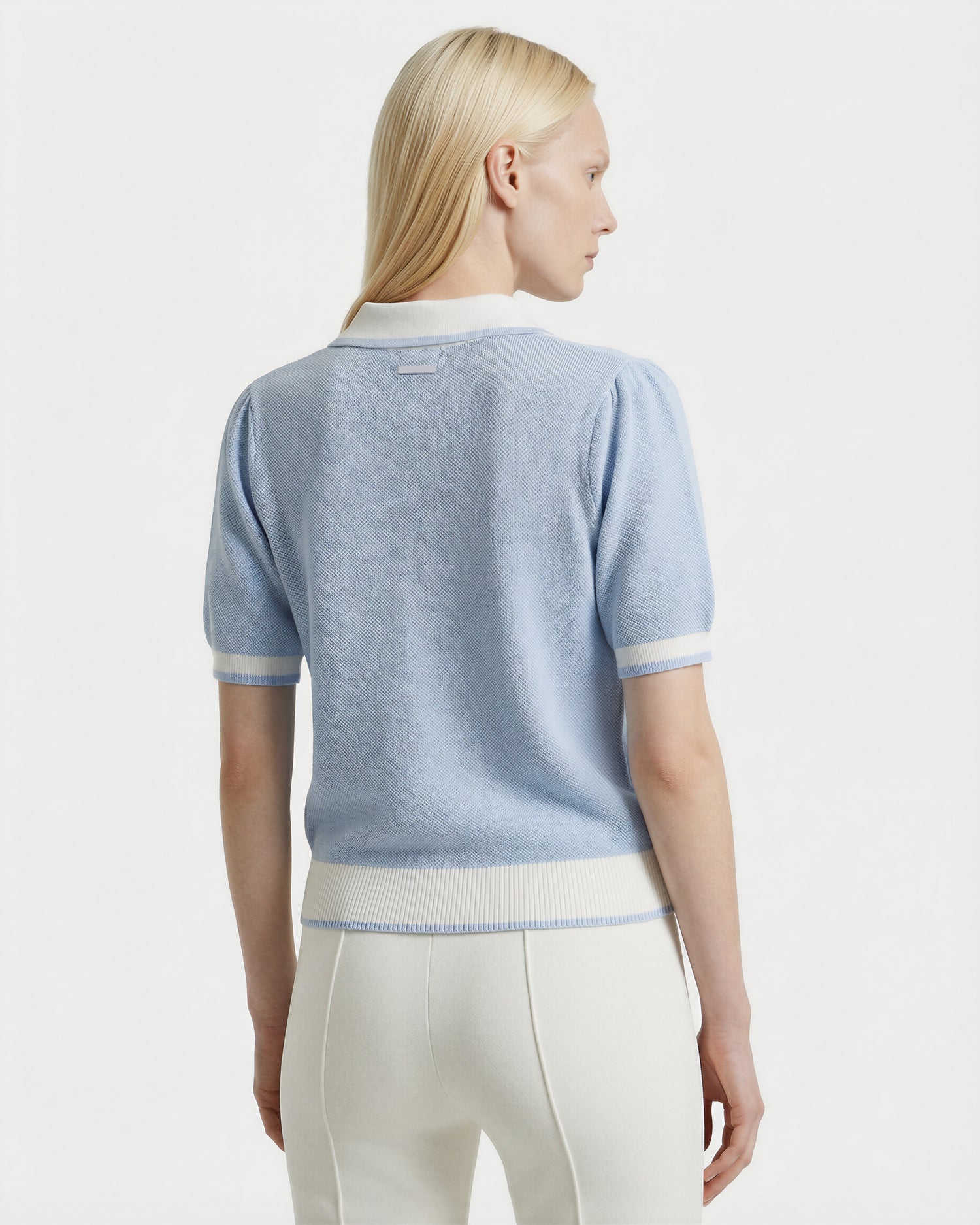 A woman with straight blonde hair, seen from behind, wears the Rudsak IRIS - 8526012 Powder Blue short-sleeve sweater and white high-waisted pants against a plain white background.