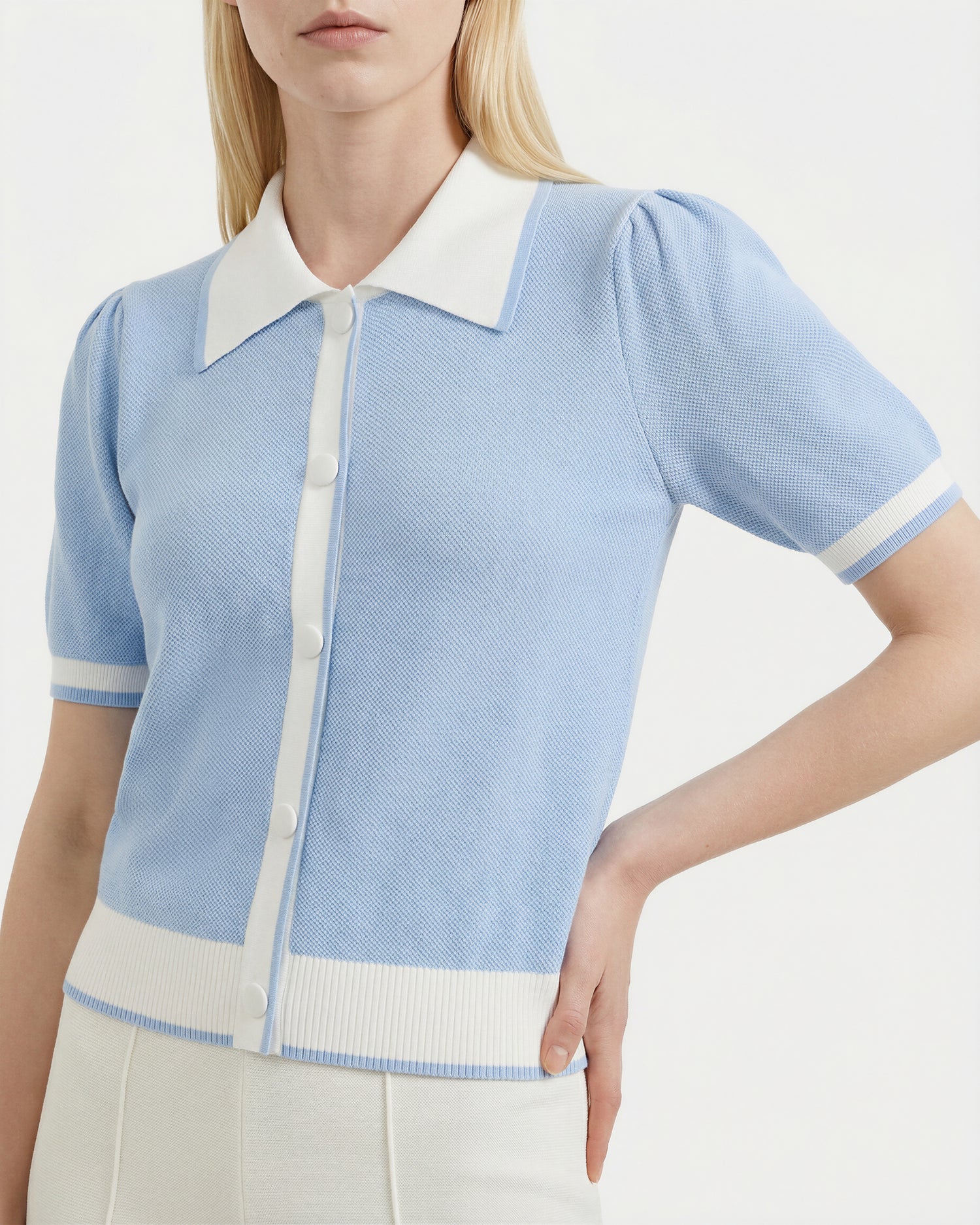 A woman wears the Rudsak IRIS - 8526012 Powder Blue knit short-sleeve sweater with contrast white collar, cuffs, and buttons, paired with high-waisted white pants. Her right hand is on her hip.