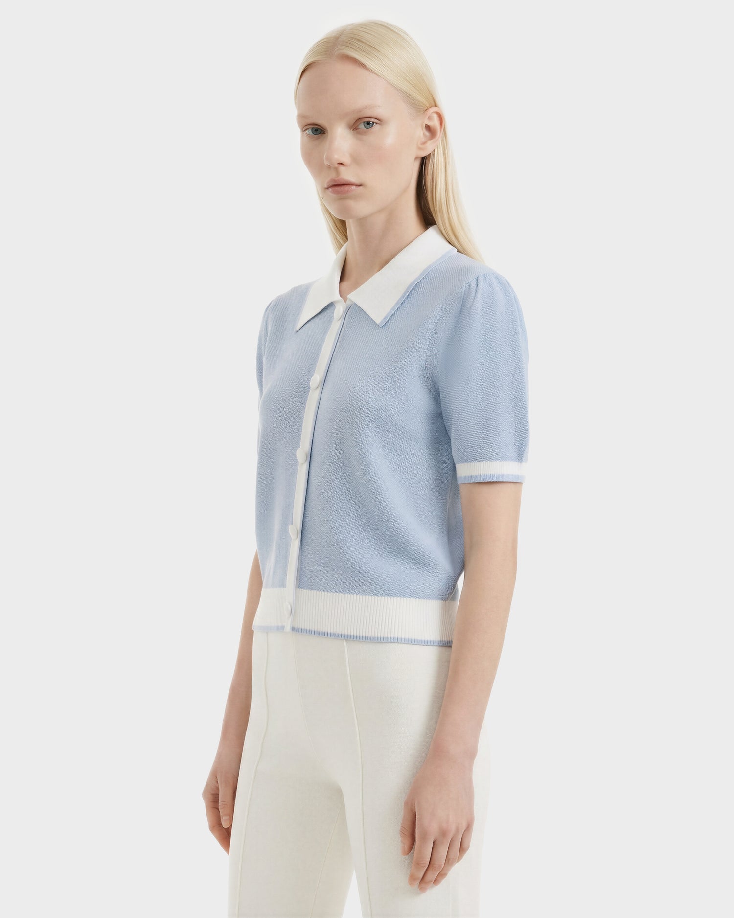 A woman with straight blonde hair models the Rudsak IRIS - 8526012 Powder Blue knit short-sleeve sweater, featuring white buttons, a contrast collar, and white trim, paired with high-waisted white pants against a plain white background.