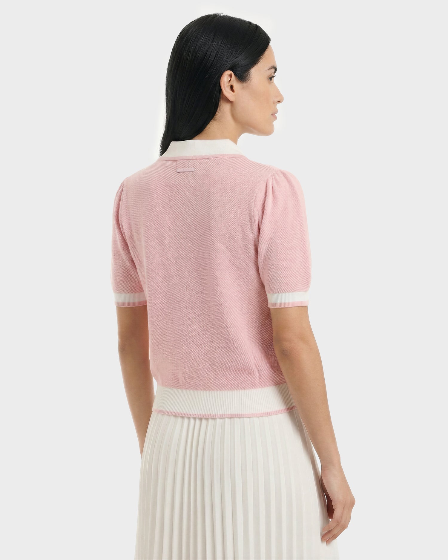 A woman with long dark hair, facing away, wears the Rudsak IRIS - 8526012 Pink knit short-sleeve sweater with white trim and a white pleated skirt against a plain light background.