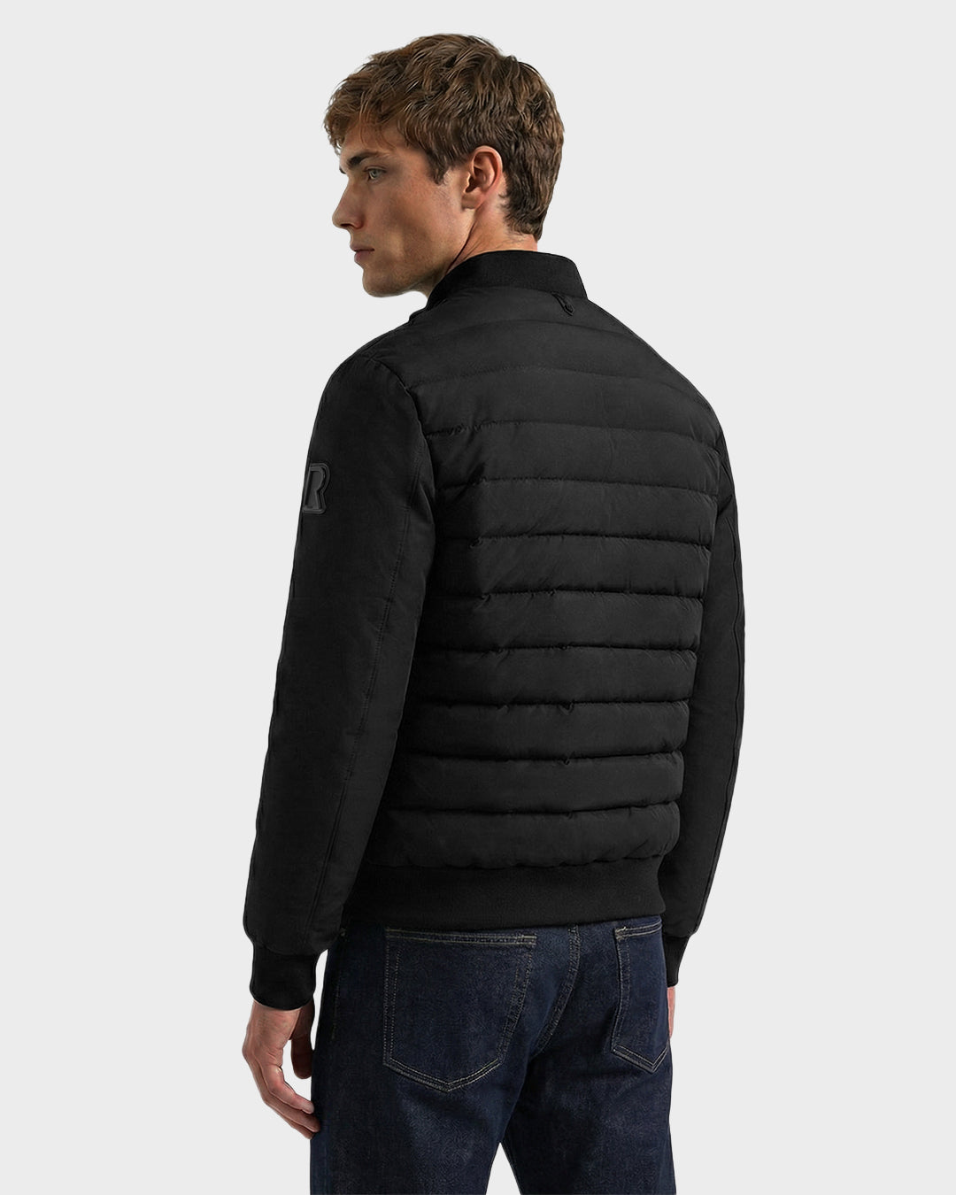 A young man with short light brown hair wears the Rudsak JIMMY - 6125723 Black men's quilted down jacket and dark blue jeans, standing against a plain light background and looking to his left.