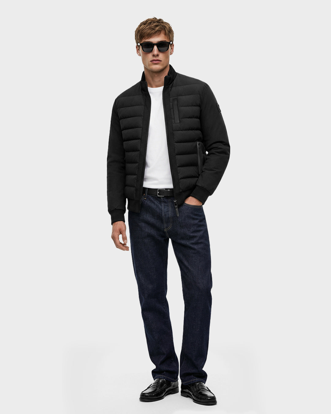 A man in dark sunglasses, a white t-shirt, dark blue jeans, black shoes, and a Rudsak JIMMY - 6125723 Black men's quilted down jacket made with RDS-certified down stands against a plain light background.