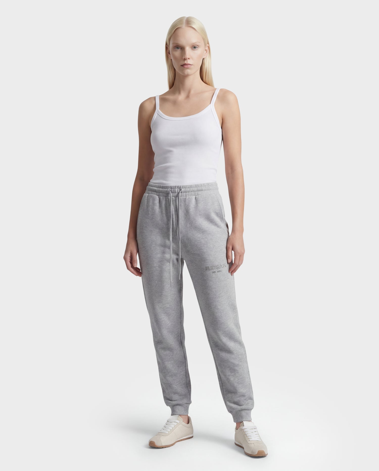 A woman stands facing forward against a plain white background, wearing the Rudsak JOSS - 9524009 Heather Grey W joggers, a white tank top, and white sneakers.