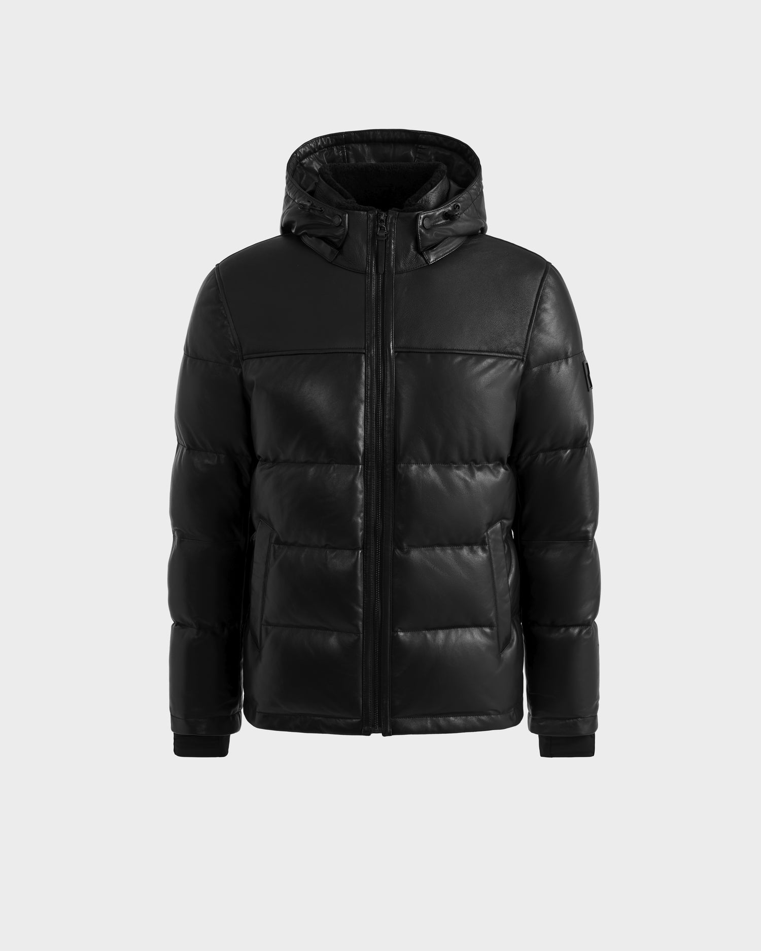 The Rudsak JAMESON - 6125678 Black men’s winter puffer jacket features a hood, front zipper, two zippered pockets, and ribbed cuffs, shown against a plain light grey background.
