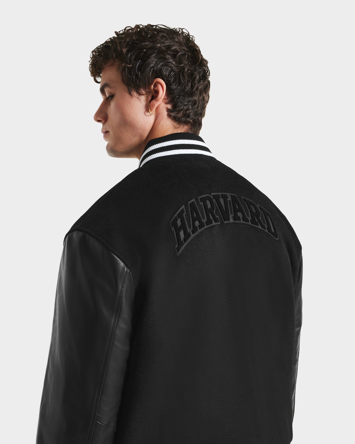 A young man with curly hair wears the limited-edition Rudsak JOHN HV - H125504 Black varsity jacket featuring leather sleeves, a white-striped collar, and HARVARD in bold arched letters across the back.