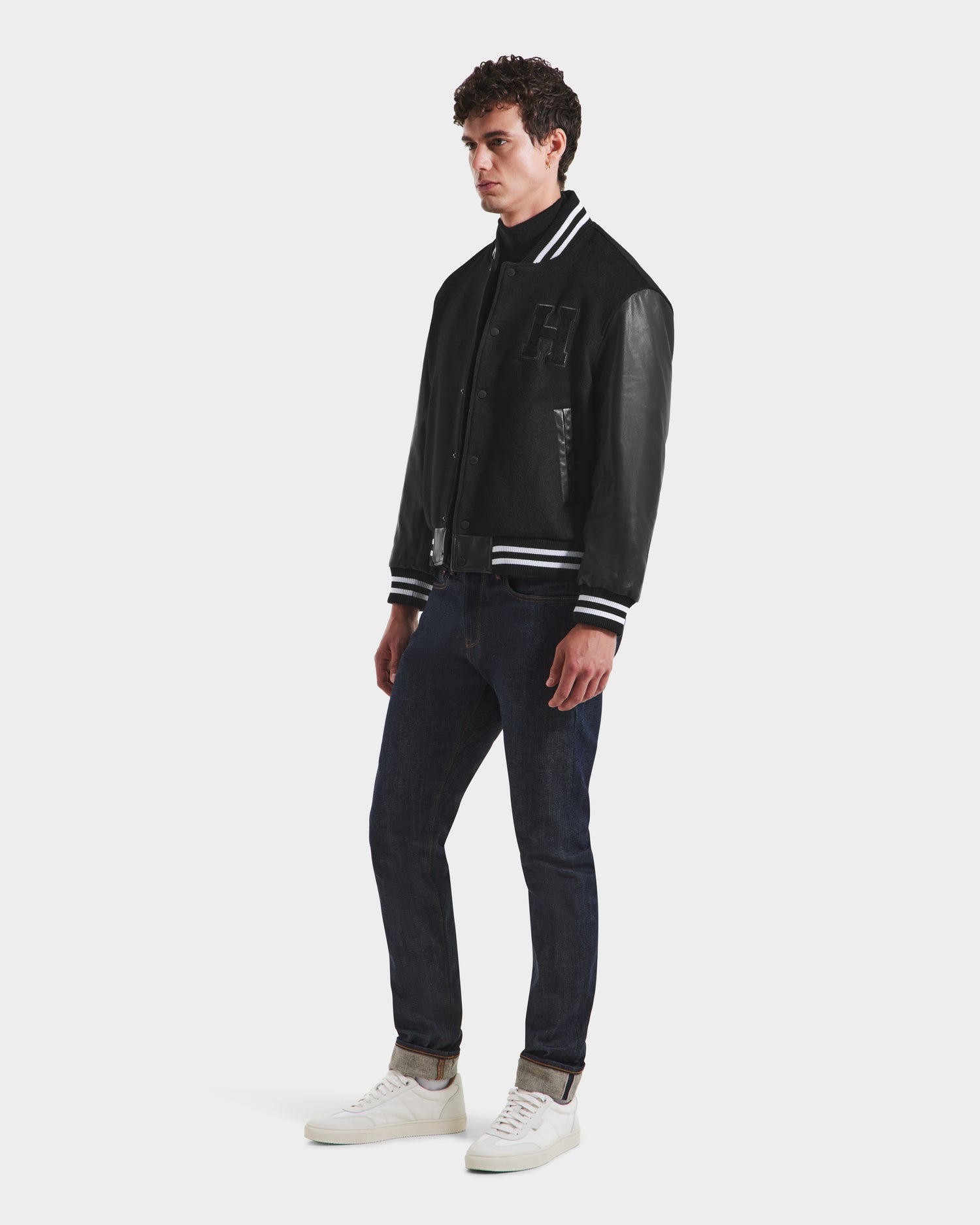 A man models the Rudsak JOHN HV - H125504 Black varsity jacket with leather sleeves, styled with dark blue jeans, a black turtleneck, and white sneakers against a plain white background.