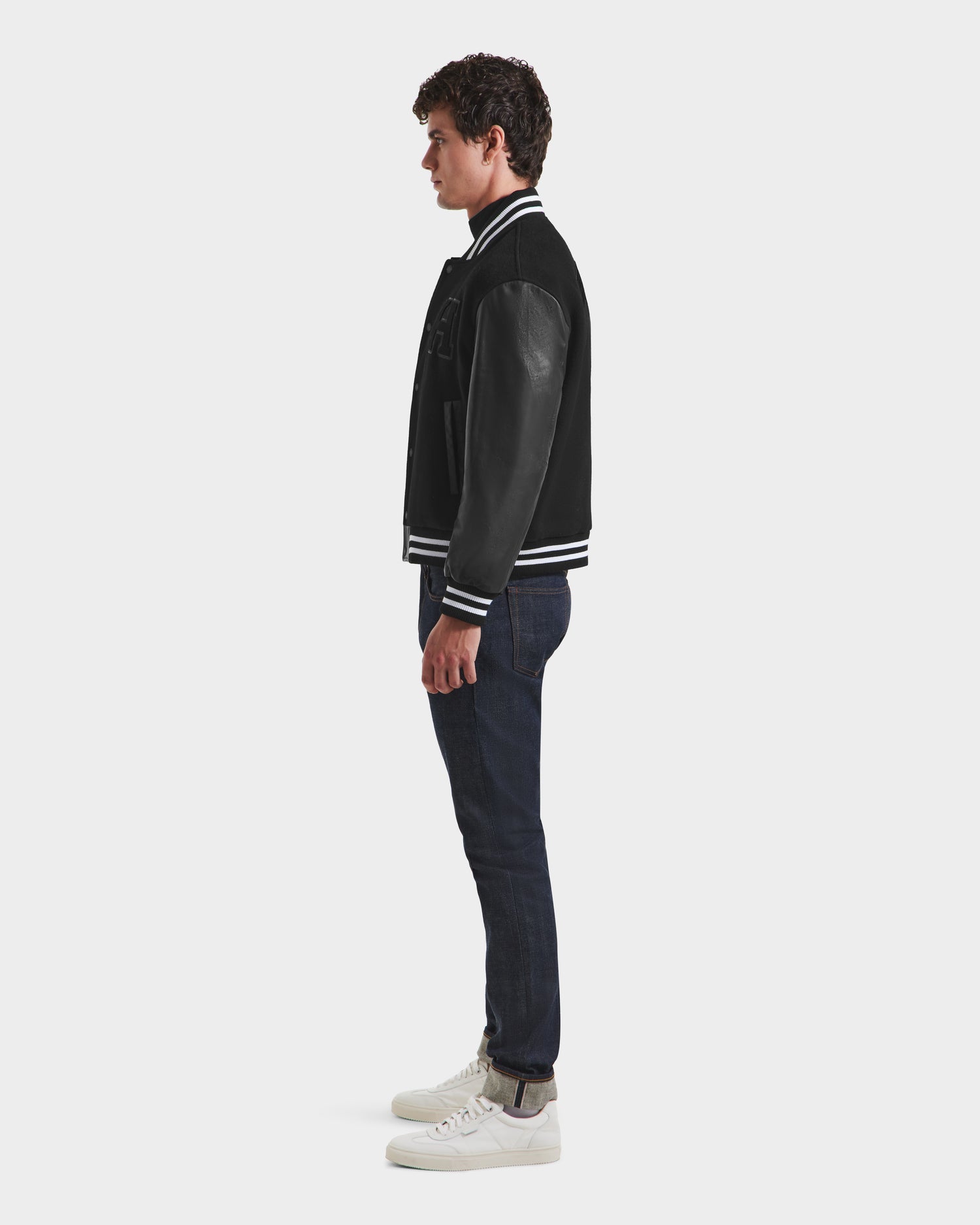 A man stands in profile against a plain background, wearing the Rudsak JOHN HV - H125504 Black varsity jacket with white trim, paired with dark blue jeans and white sneakers.