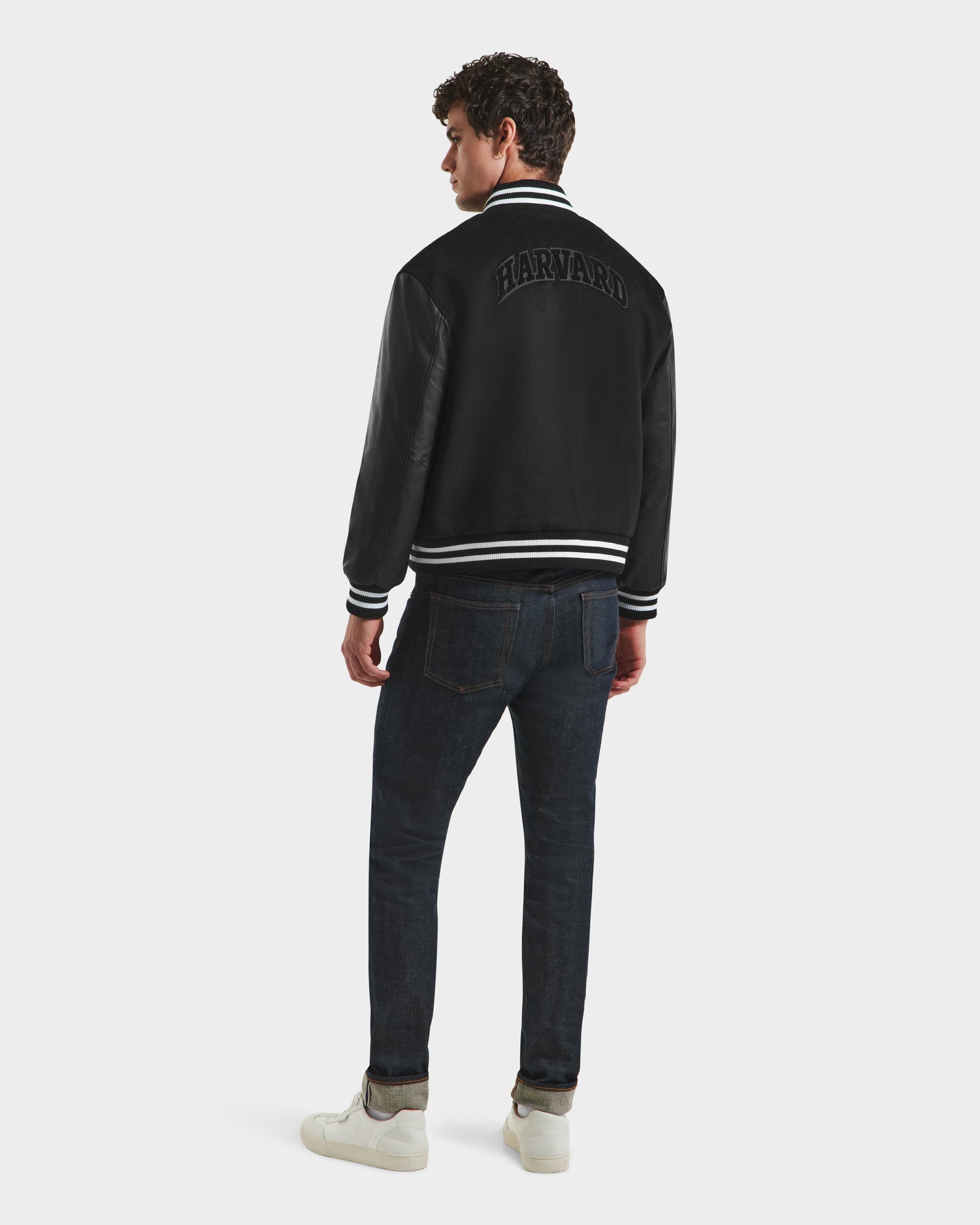 A man faces away, wearing Rudsak's JOHN HV - H125504 Black varsity jacket, paired with dark jeans and white sneakers against a plain light background.