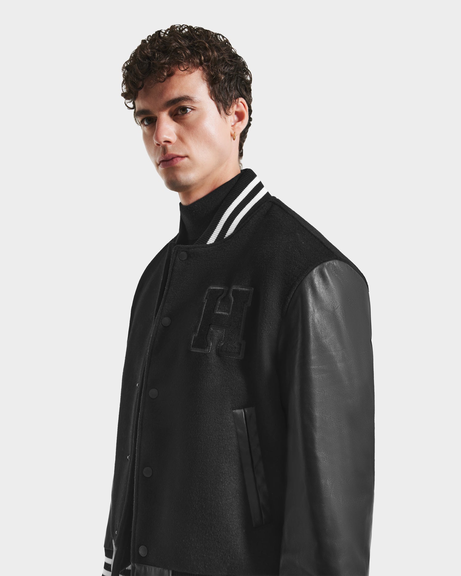 A young man with short curly hair wears the Rudsak JOHN HV - H125504 Black varsity jacket. It features leather sleeves and a bold H on the chest. He stands with a serious expression against a plain white background.