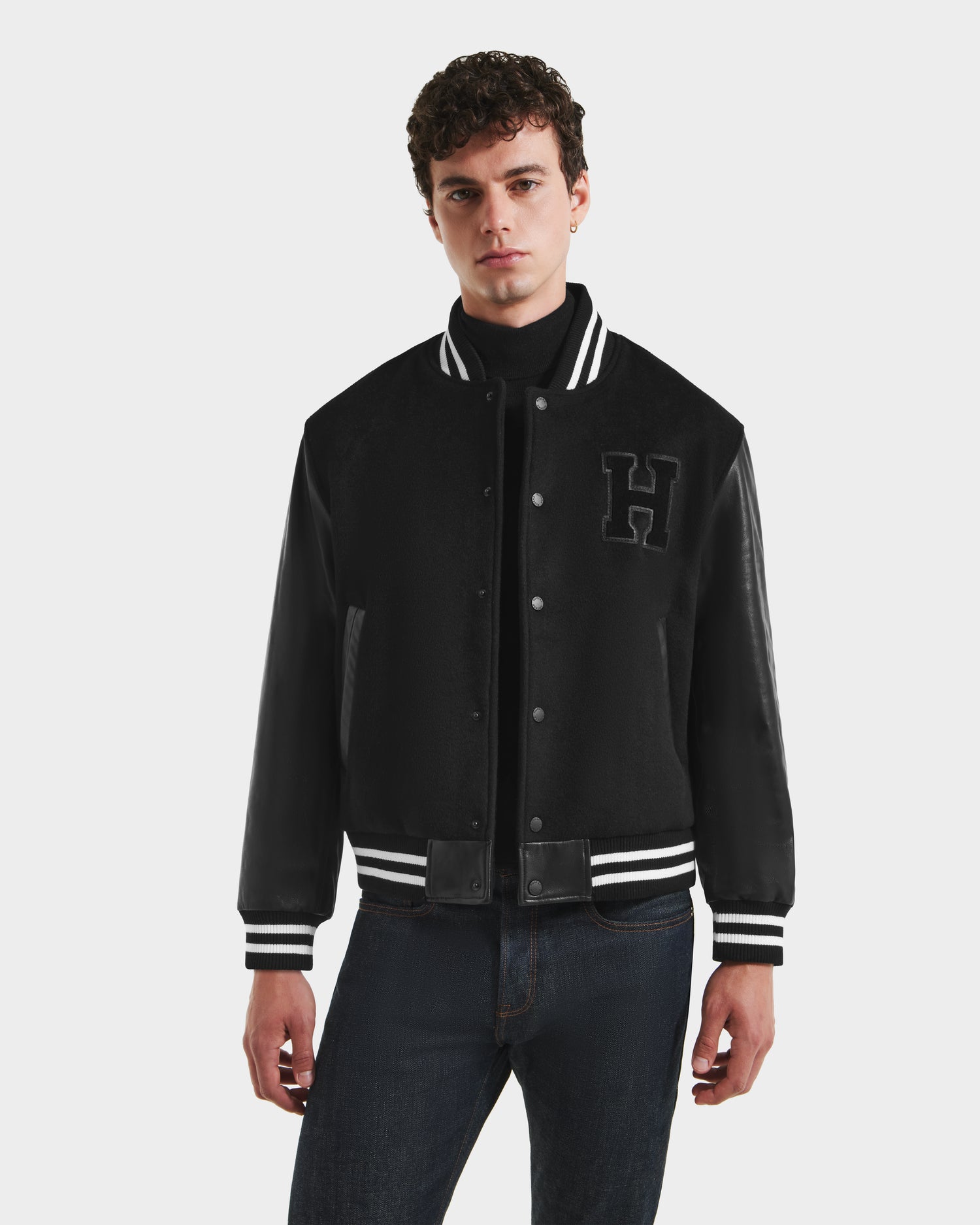 A young man wears the Rudsak JOHN HV - H125504 Black varsity jacket with white striped cuffs and collar and a large "H" patch on the left chest, standing against a white background.
