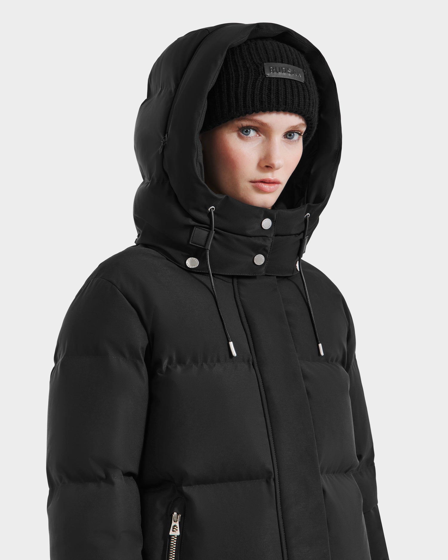 A woman, slightly turned to the side, wears the Rudsak JOON HV - H125506 Black women’s heritage puffer jacket with a hood and visible drawstrings, plus a black knit beanie. The premium down jacket has zippered pockets and is set against a plain white background.