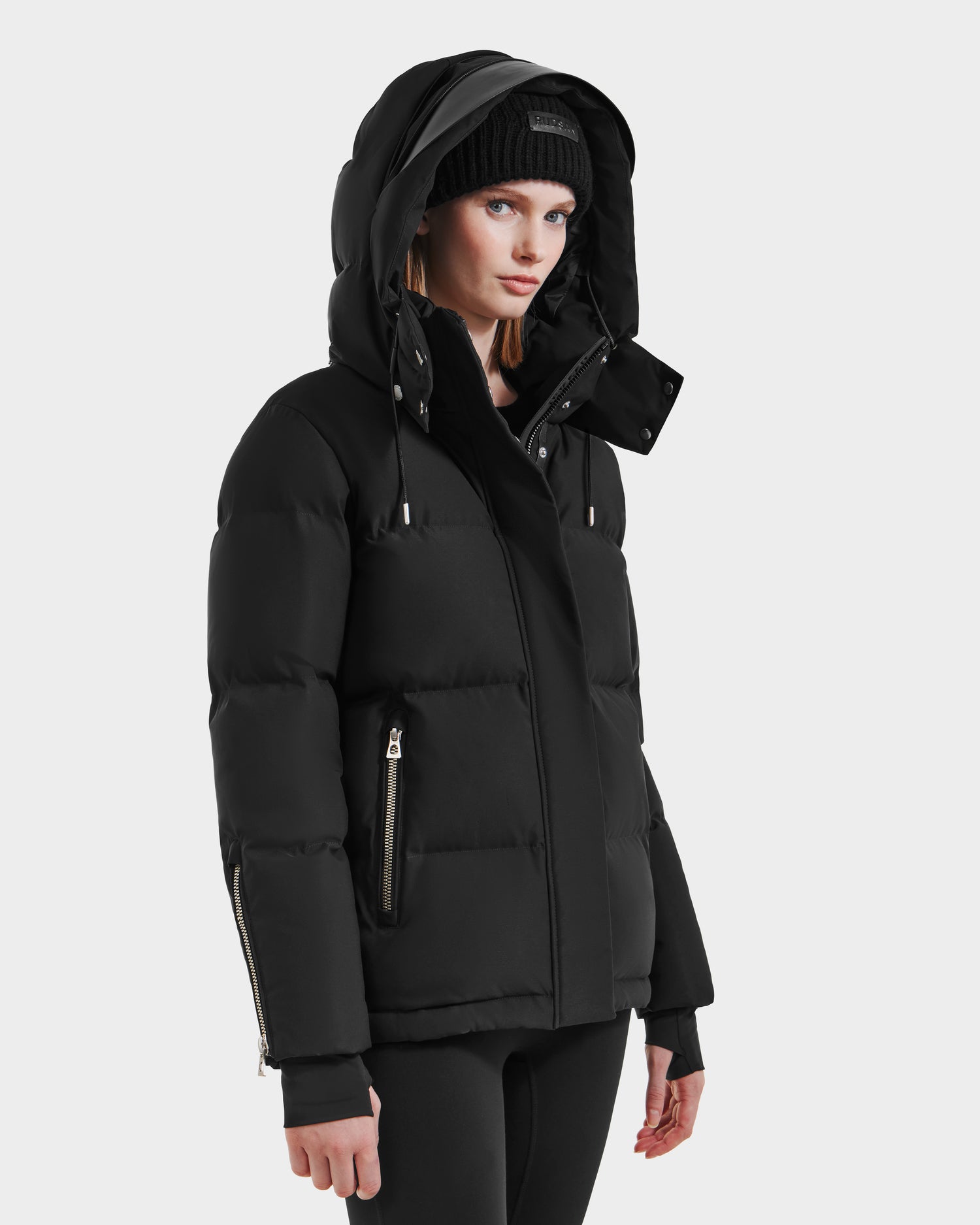 A woman models the Rudsak JOON HV - H125506 Black heritage puffer with hood and zippered pockets, styled with a black knit hat and pants against a plain light background.