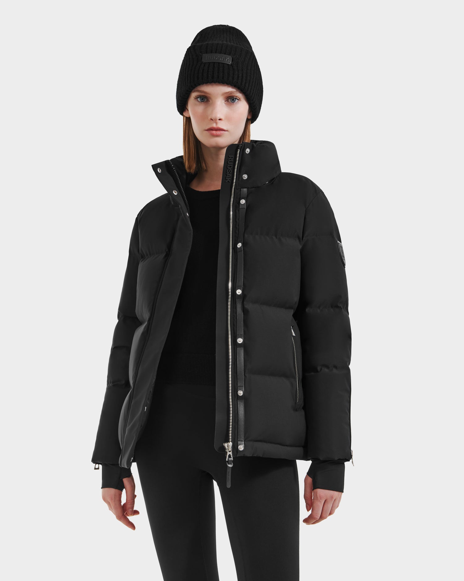 A woman stands facing forward against a plain light background, wearing Rudsak’s JOON HV - H125506 Black premium down women’s heritage puffer with silver zippers, paired with a black beanie, sweater, and pants.