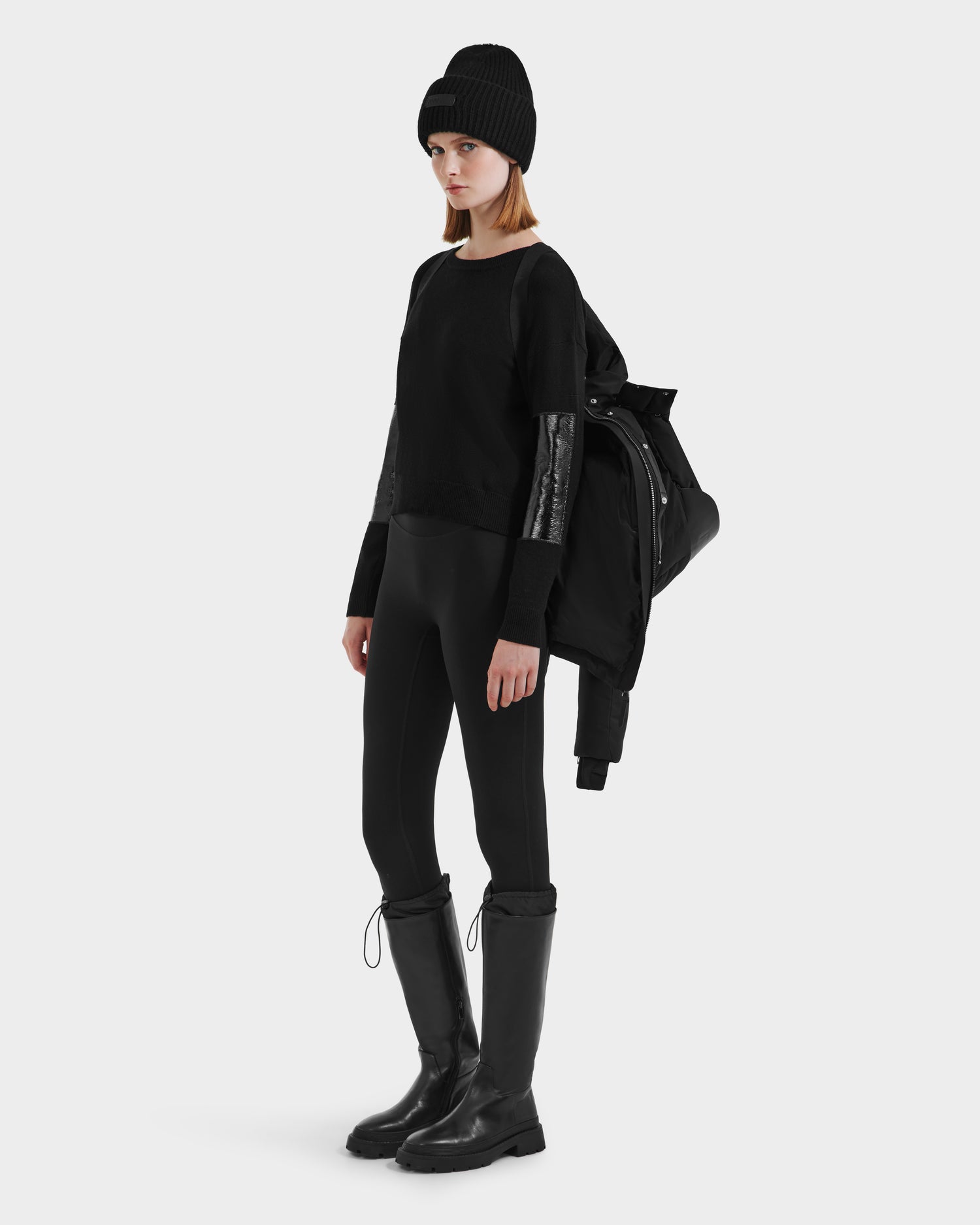 Wearing black leggings, a black sweater with shiny sleeves, knee-high boots, and a black knit beanie, a person holds the Rudsak JOON HV - H125506 Black premium down jacket over one shoulder against a plain light background.