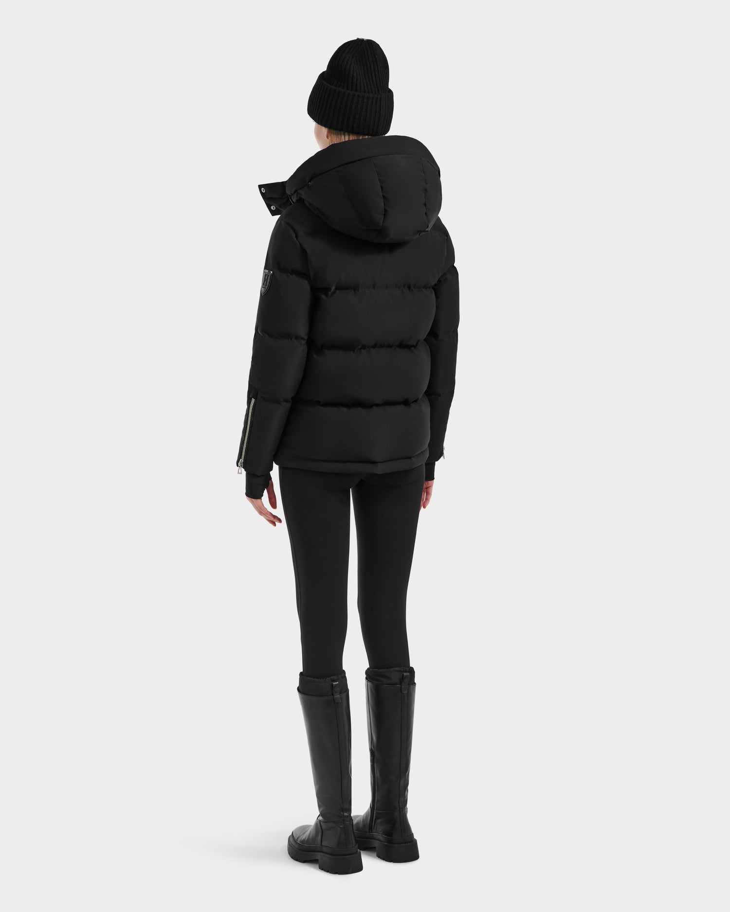 A person wearing the Rudsak JOON HV - H125506 Black premium down women’s heritage puffer jacket, a black beanie, black leggings, and knee-high boots stands facing away against a plain light background.