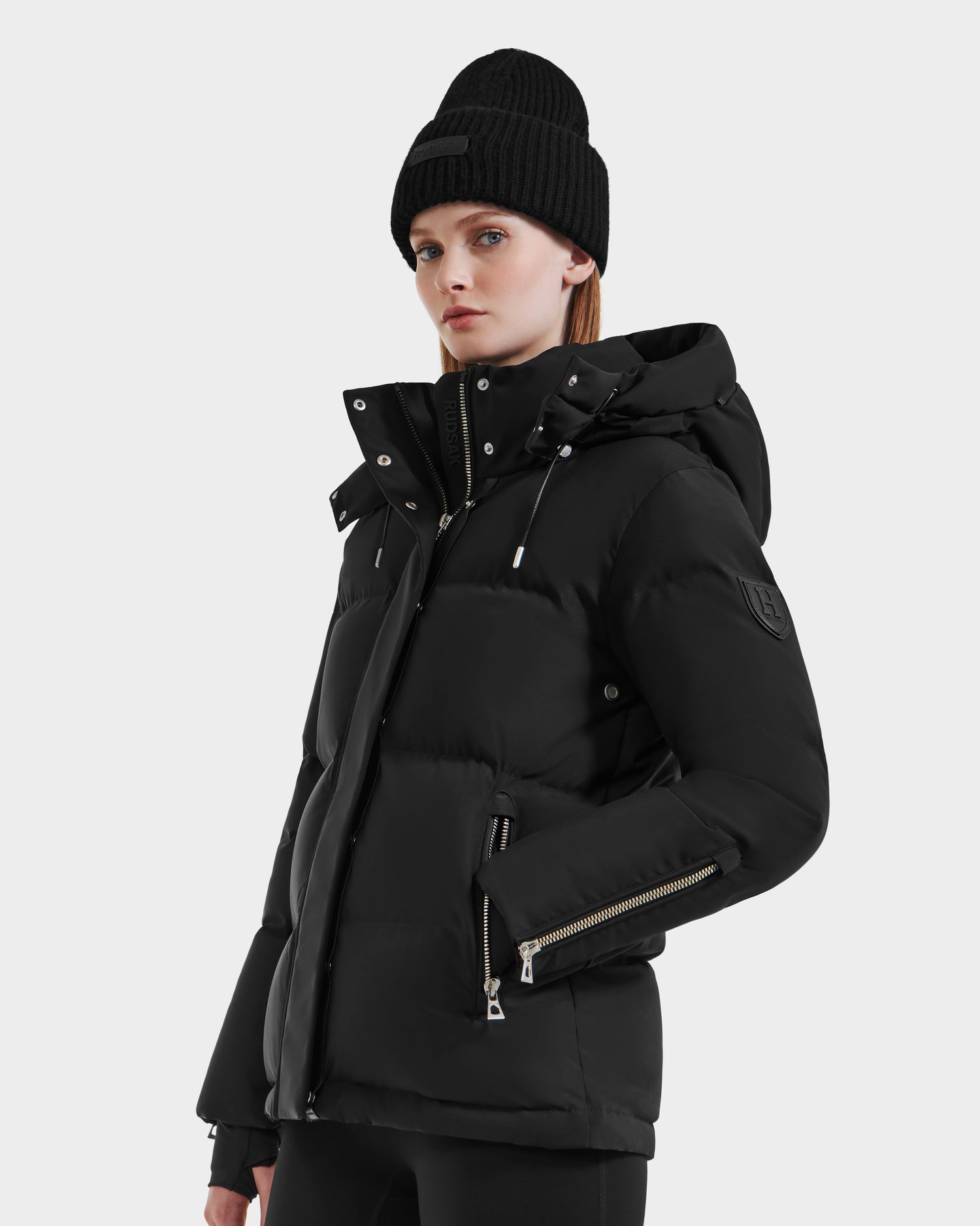 A woman wears the Rudsak JOON HV - H125506 Black heritage puffer with a hood and zippered pockets, styled with a black knit beanie. She stands against a plain background with a neutral expression.