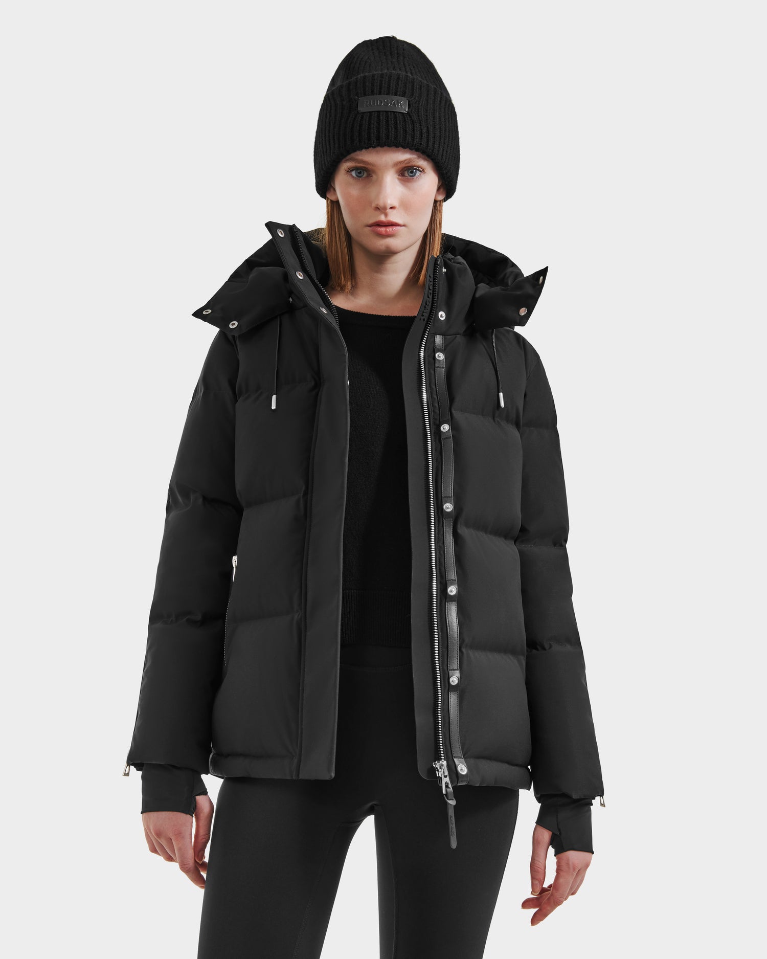 A woman wearing the Rudsak JOON HV - H125506 Black premium down puffer jacket, a black knit beanie, and black leggings stands facing forward against a plain white background.