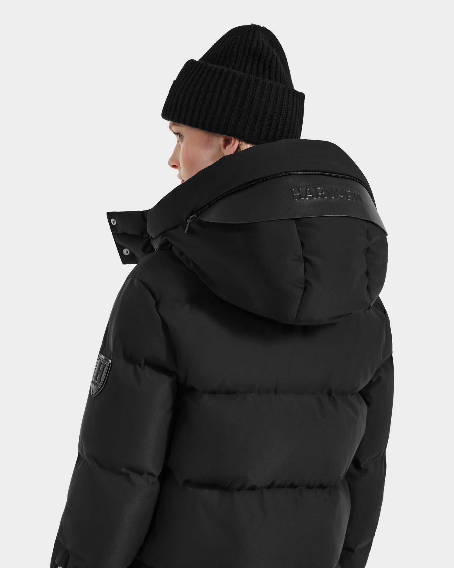 A person in a Rudsak JOON HV - H125506 Black women’s heritage puffer with premium down, embossed hood logo, and sleeve patch faces away on a light gray background, styled with a black knit beanie.