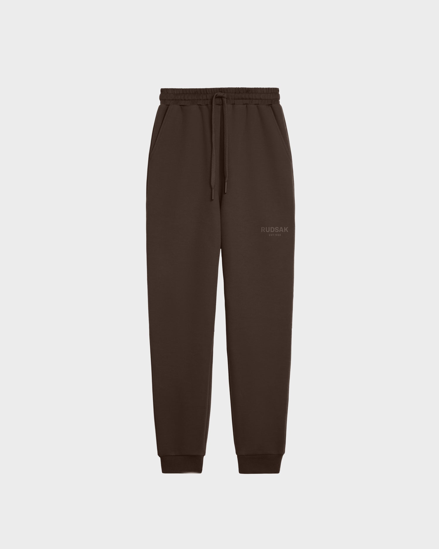 The JOSS - 9524009 Espresso W by Rudsak is a dark brown unisex jogger made of premium cotton with an elastic waistband, drawstring, side pockets, ribbed cuffs, and a subtle Rudsak logo on the left thigh. Displayed on a plain white background.