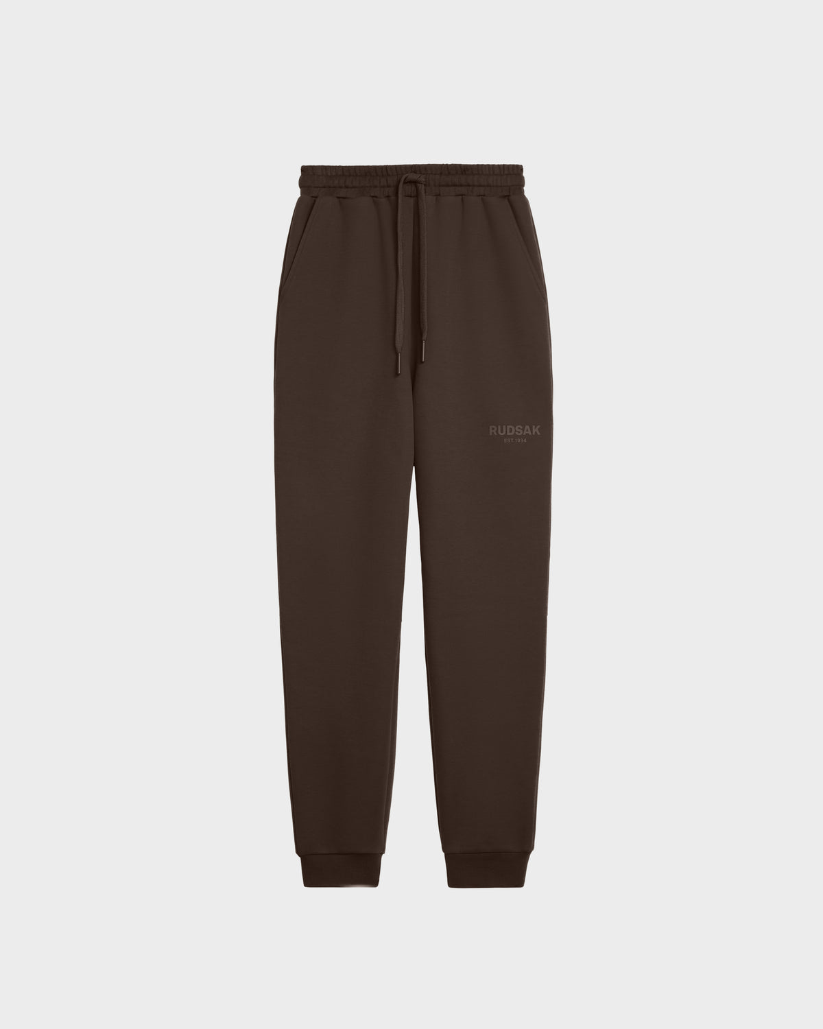 The JOSS - 9524009 Espresso W by Rudsak is a dark brown unisex jogger made of premium cotton with an elastic waistband, drawstring, side pockets, ribbed cuffs, and a subtle Rudsak logo on the left thigh. Displayed on a plain white background.