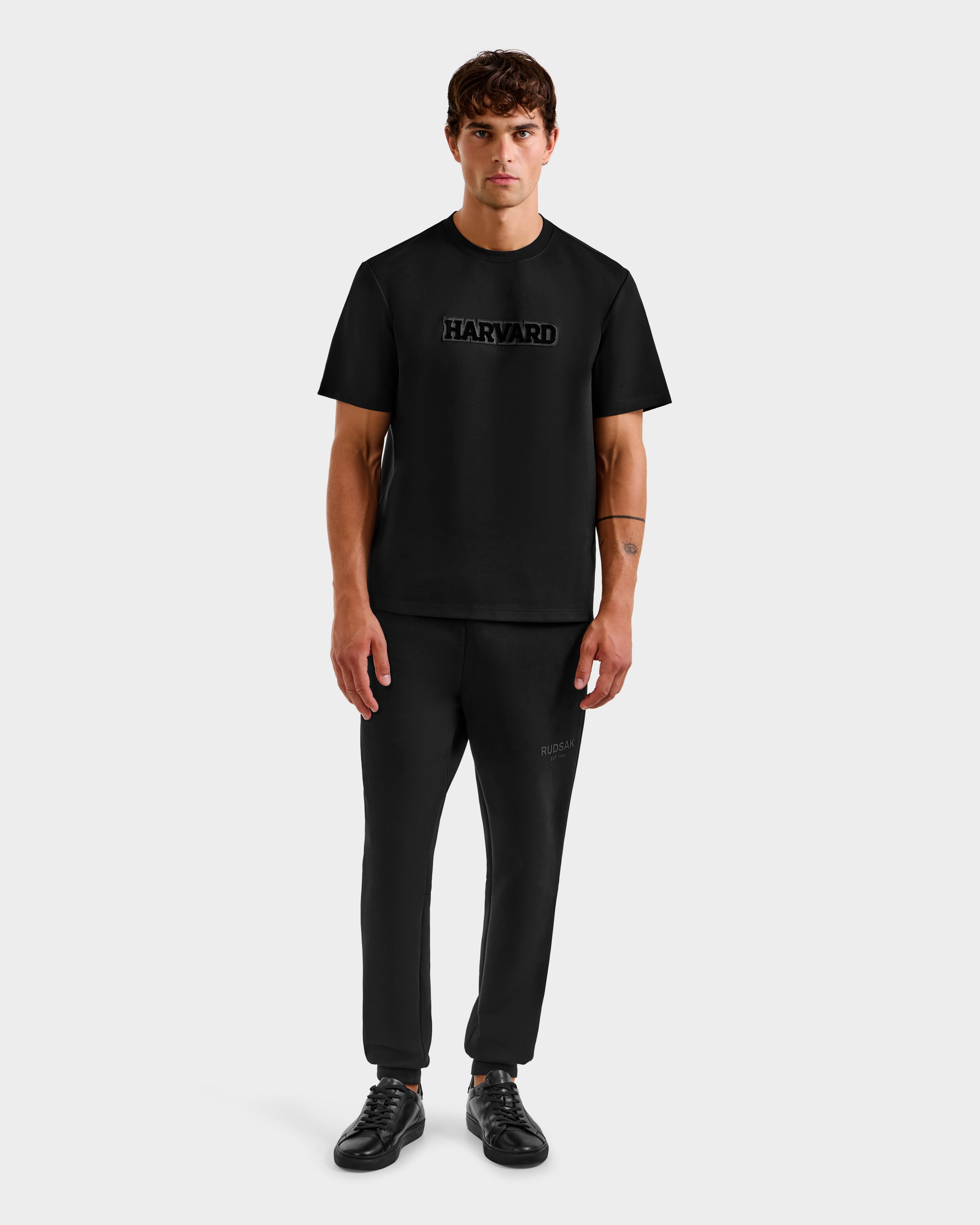 A young man stands facing forward in a limited-edition Harvard t-shirt, black pants, and Rudsak KARTER B HV - H525508 Black sneakers against a plain light gray background—capturing classic Ivy League style.