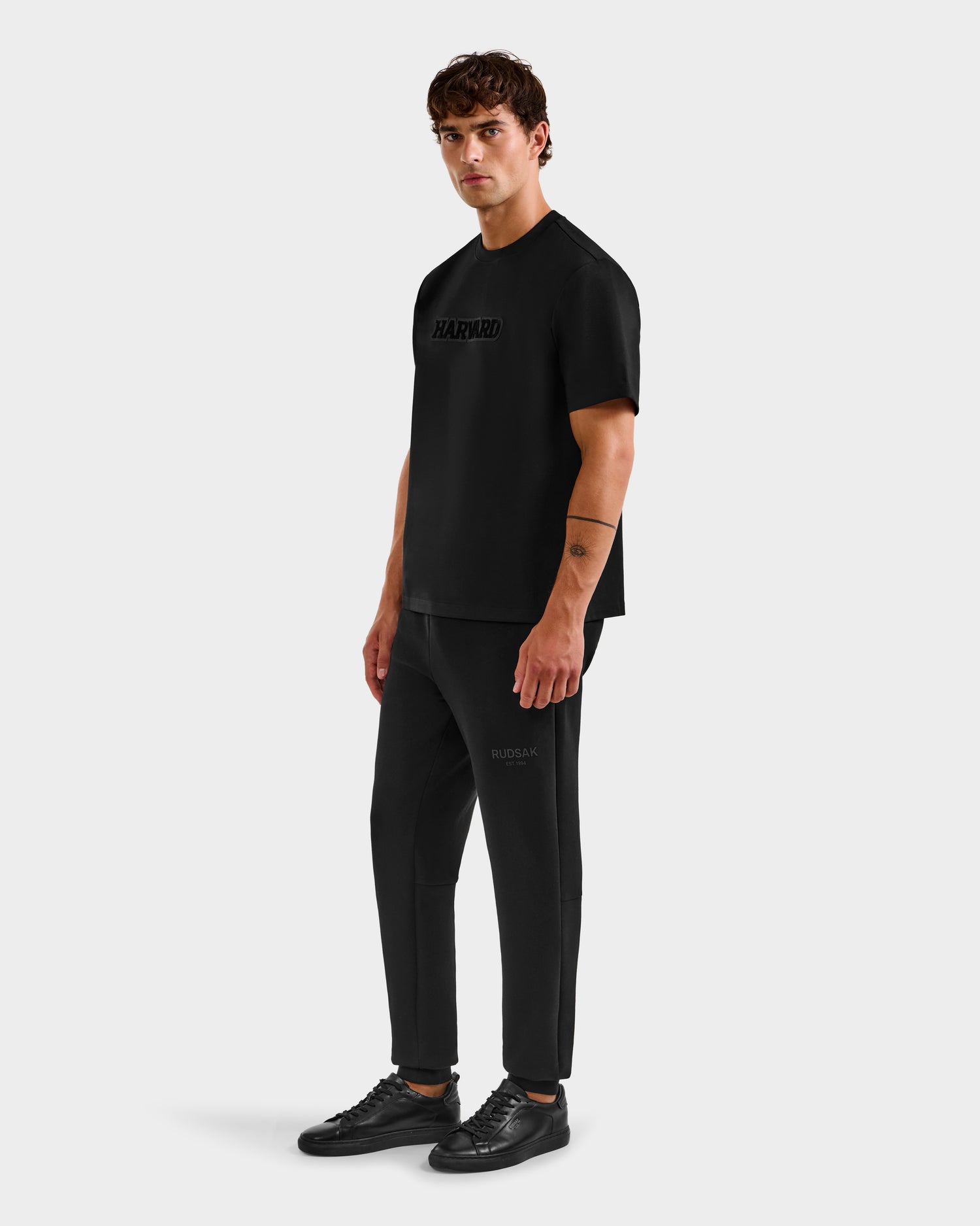 A man stands against a plain background wearing the limited-edition Harvard t-shirt, black pants, and Rudsak KARTER B HV - H525508 Black sneakers. He faces slightly to the side with a neutral expression, projecting classic Ivy League style.