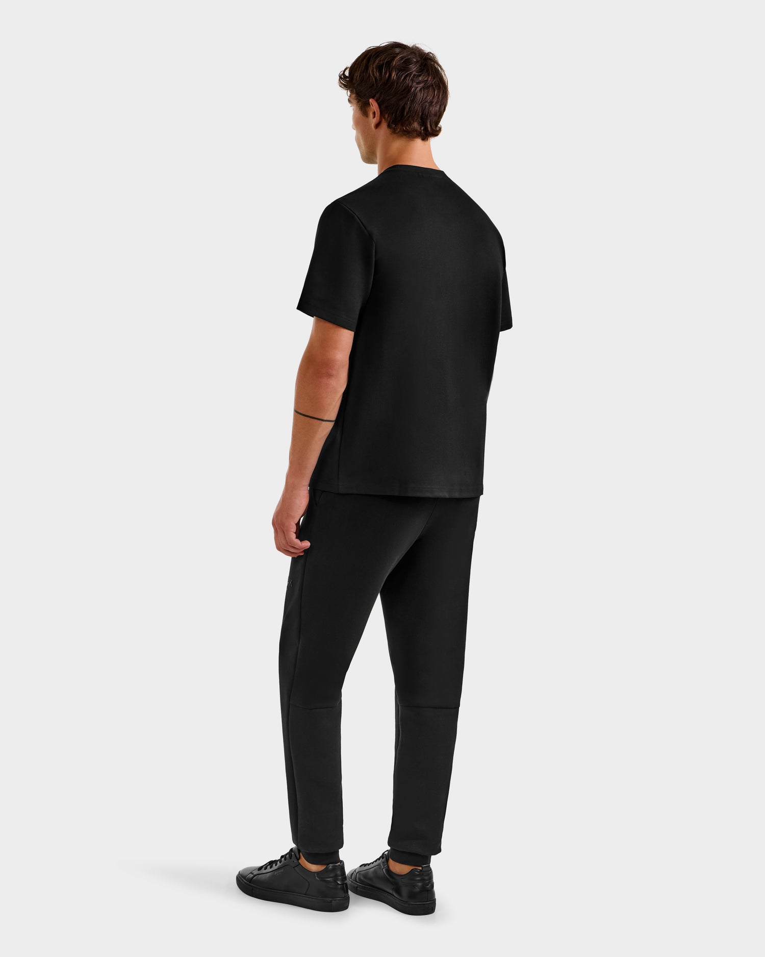 A man facing away from the camera wears Rudsak's KARTER B HV - H525508 Black t-shirt, paired with black pants and sneakers, set against a plain light gray background.