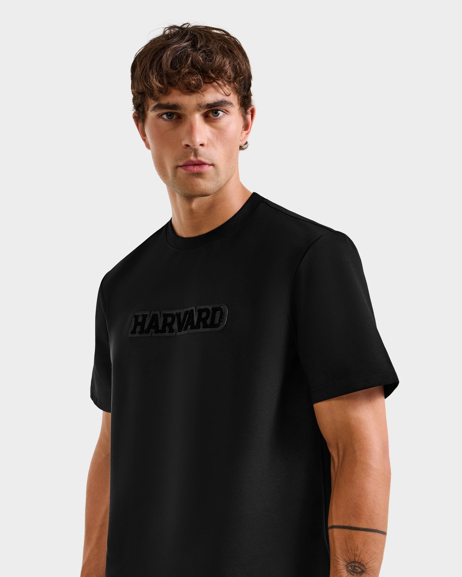 A young man with short brown hair models the Rudsak KARTER B HV - H525508 Black t-shirt, a limited-edition piece with raised black letters. He stands against a plain light background, capturing timeless Ivy League style.