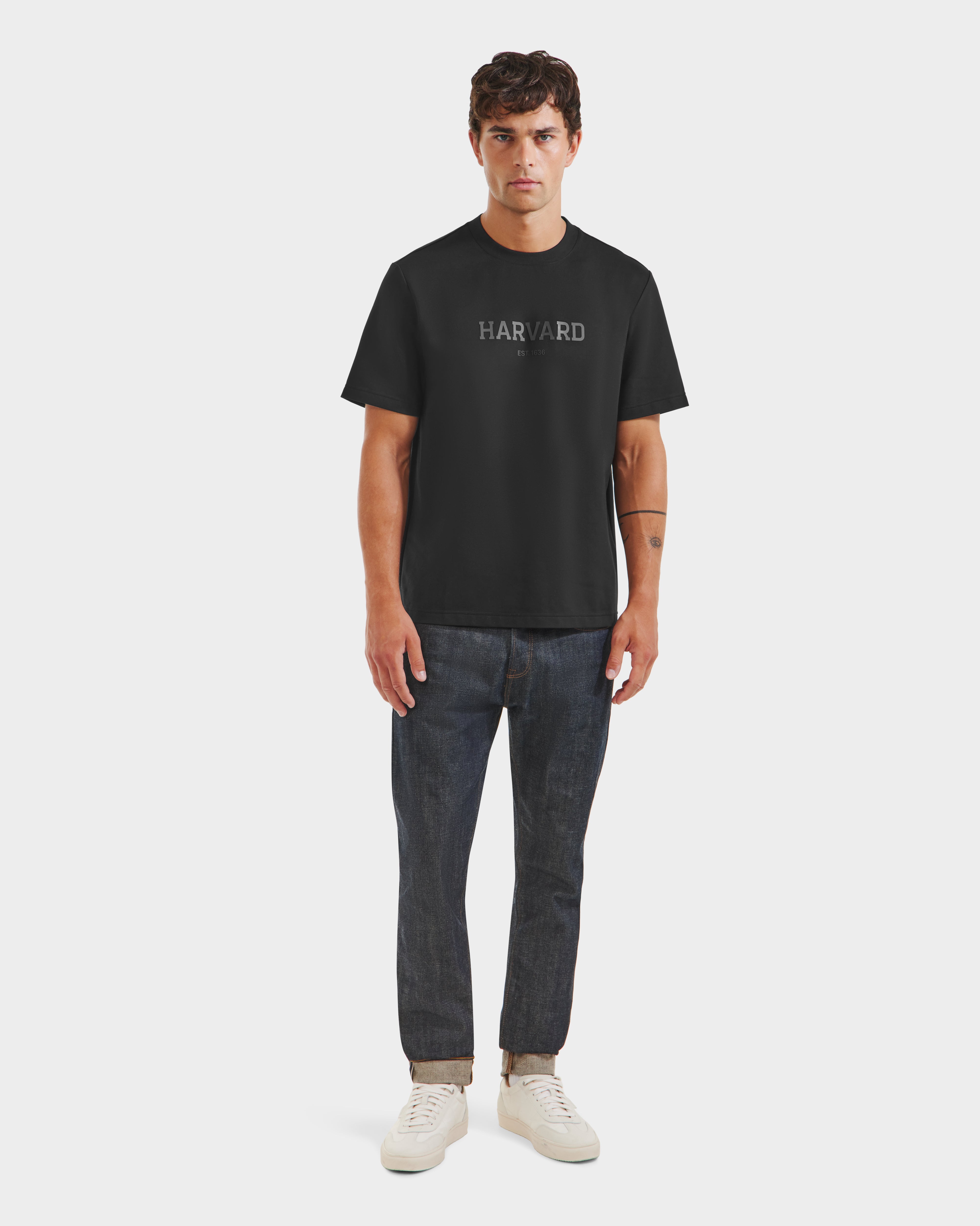 A man stands facing forward in limited edition Harvard T-shirt, dark cuffed jeans, and black Rudsak KARTER R HV - H525507 sneakers against a plain light background.