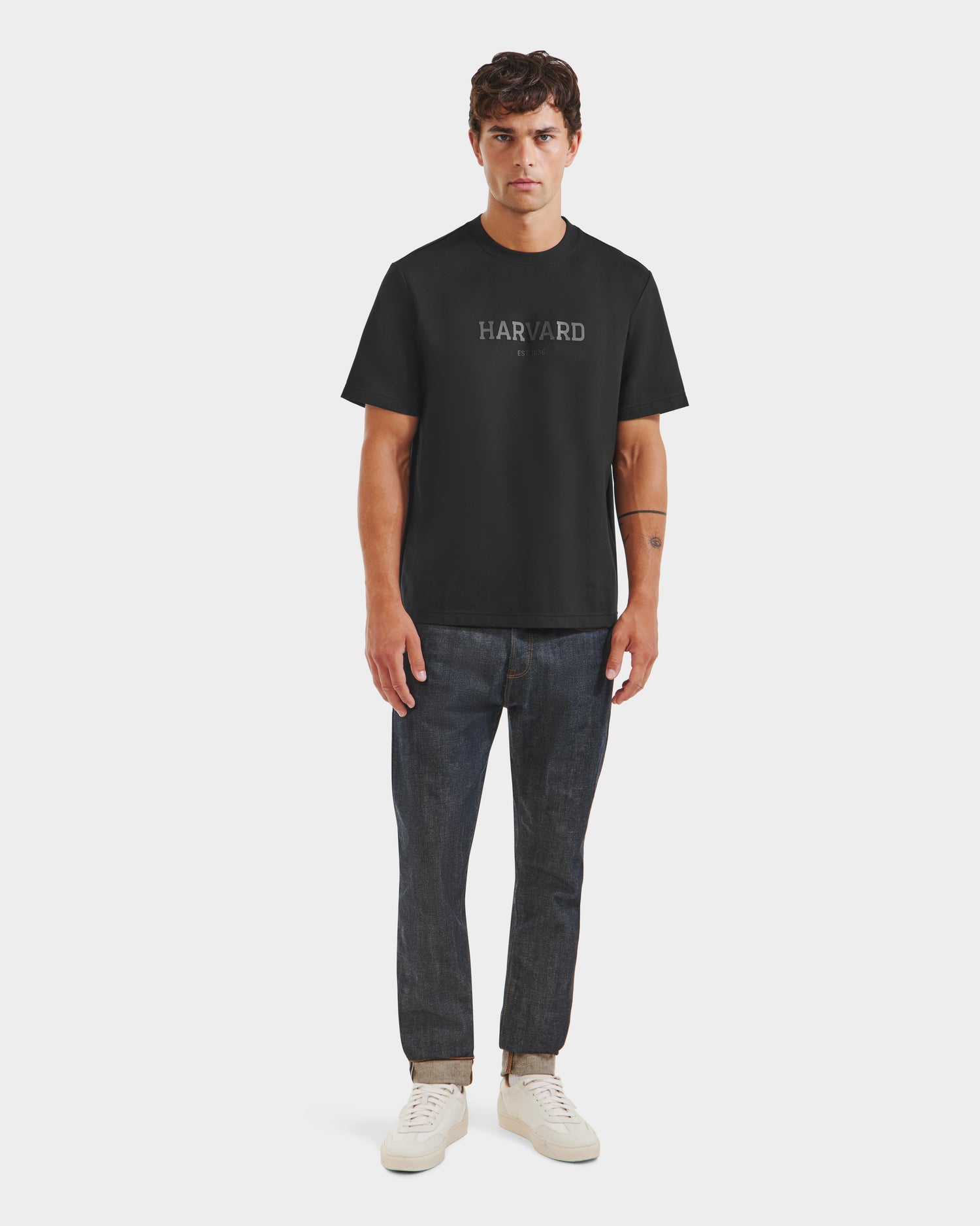 A man stands facing forward in limited edition Harvard T-shirt, dark cuffed jeans, and black Rudsak KARTER R HV - H525507 sneakers against a plain light background.
