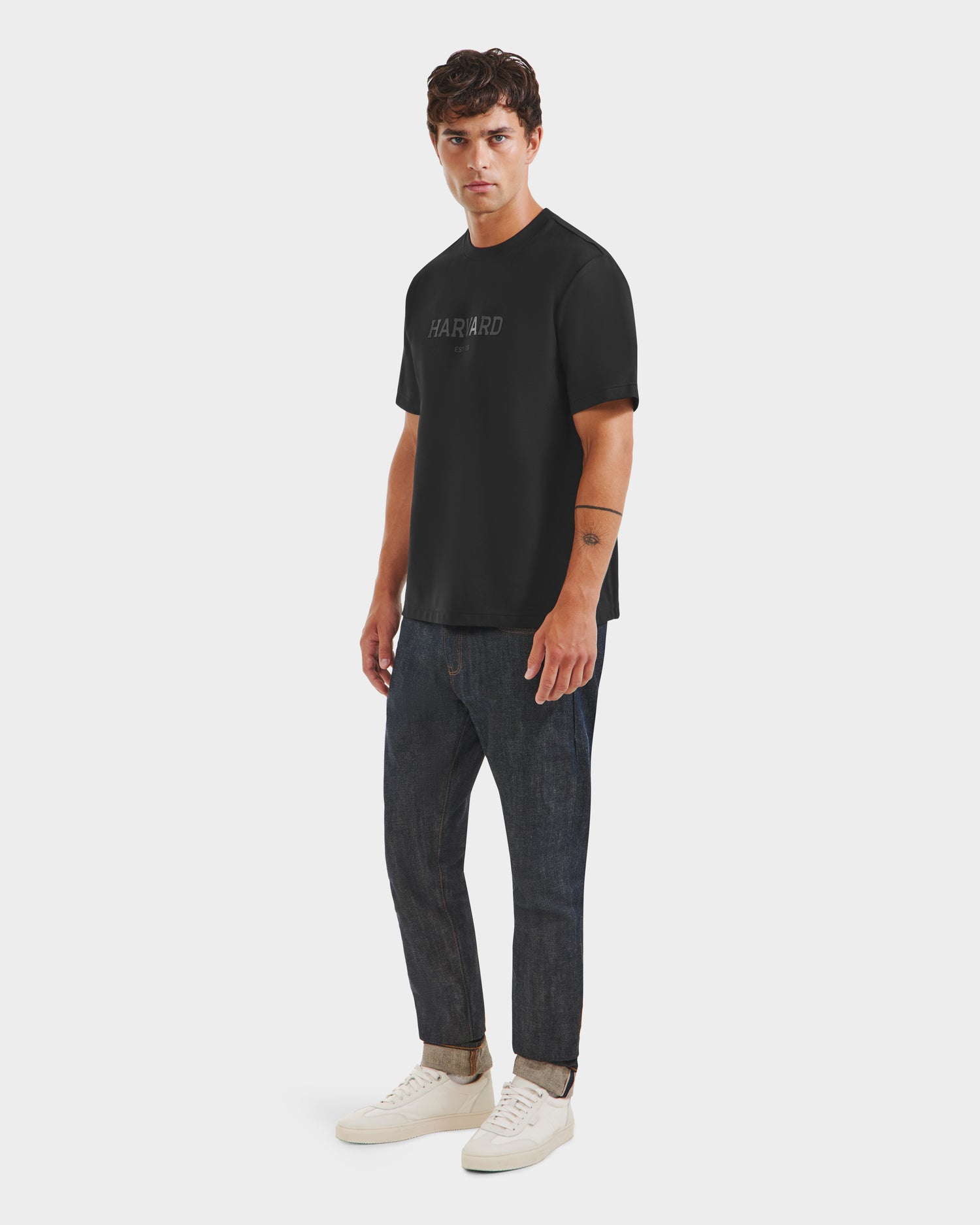 A man faces forward in a KARTER R HV - H525507 Black T-shirt by Rudsak, styled with dark rolled-cuff jeans and white sneakers. He has short dark hair and a neutral expression, showcasing streetwear style on a plain white background.