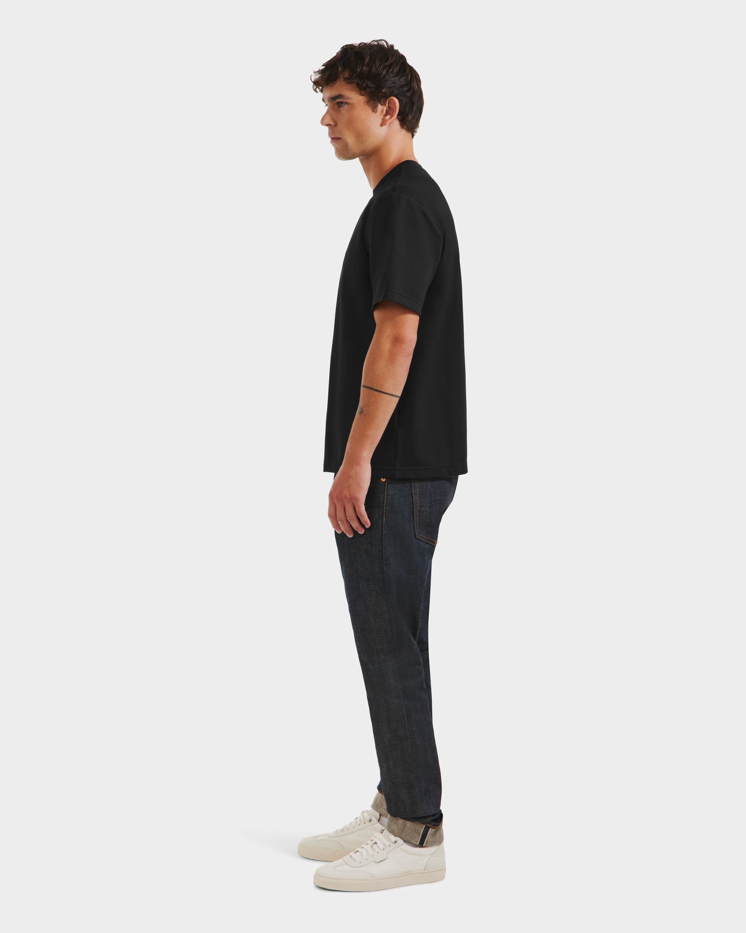 A man stands in profile against a white background, wearing the limited edition KARTER R HV - H525507 Black T-shirt by Rudsak with dark jeans and white sneakers—effortlessly showcasing cool streetwear style.